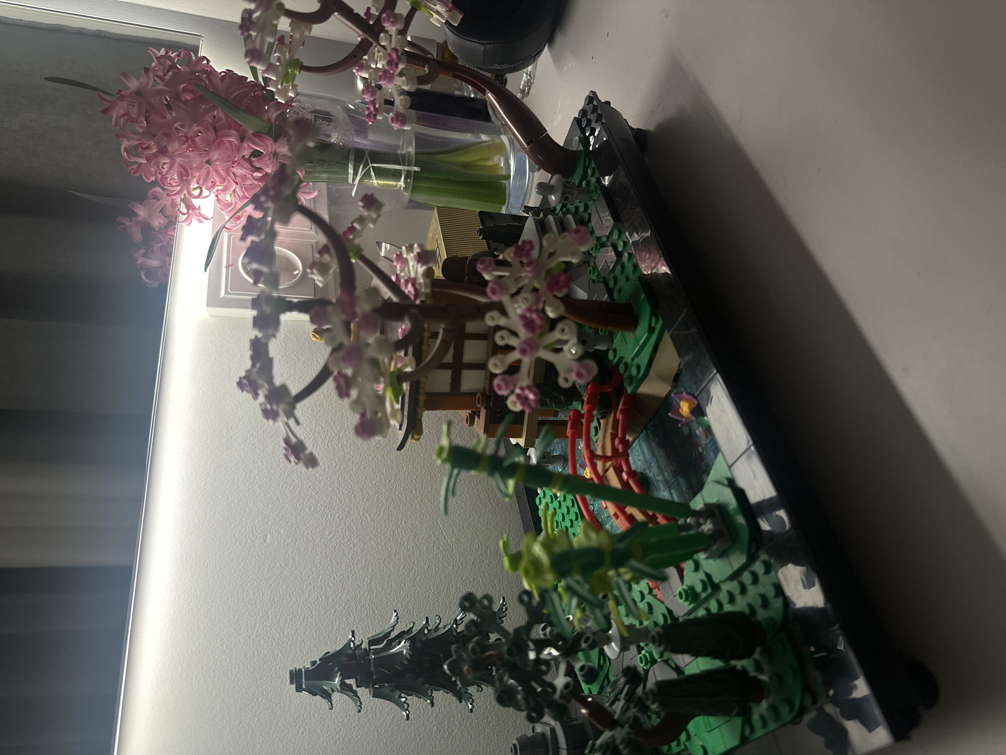 A Lego bonsai tree is in a black pot on a white surface. A pink hyacinth flower is next to the tree in a glass vase with cut celery stalks. A light fixture is on the wall behind the hyacinth flower. The photograph was likely taken indoors. Yerevan, Armenia is not evident in the image.