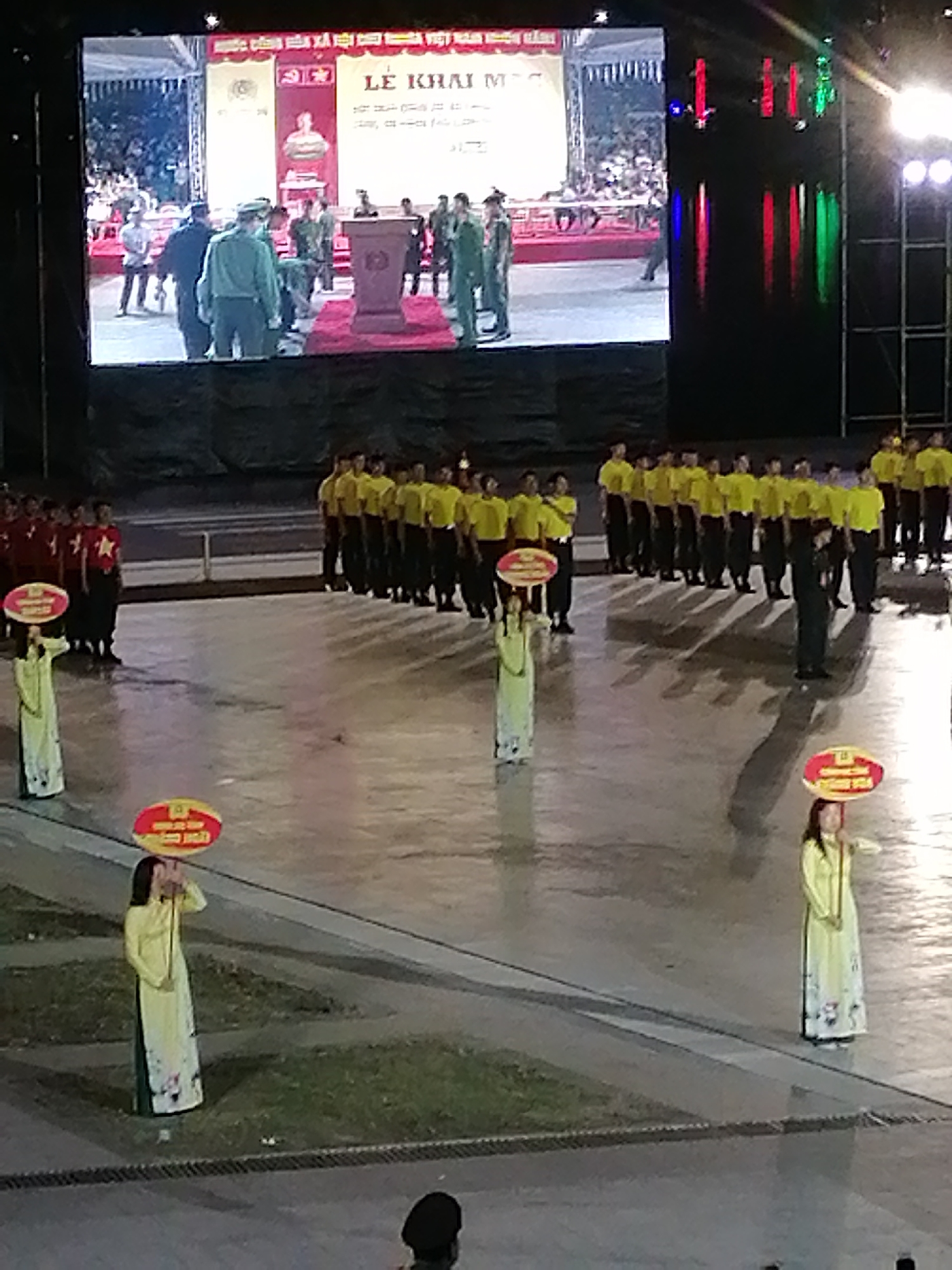 The image captures a wide-angle view of an outdoor event, likely a ceremony or performance, taking place at night in Da Lat, Vietnam. A large LED screen dominates the background, displaying a scene with people in military-style uniforms and a podium. In the foreground, a group of individuals in matching yellow shirts stand in formation, facing away from the camera. To the left of this group, several individuals in red shirts also stand in formation. Two women in traditional Vietnamese long dresses, known as "áo dài," are positioned in the foreground, each holding a circular sign. The ground is paved and appears wet, possibly from recent rain, reflecting the lights from the event. The overall atmosphere suggests a formal occasion with a public audience, indicated by the rows of people and the large display screen.