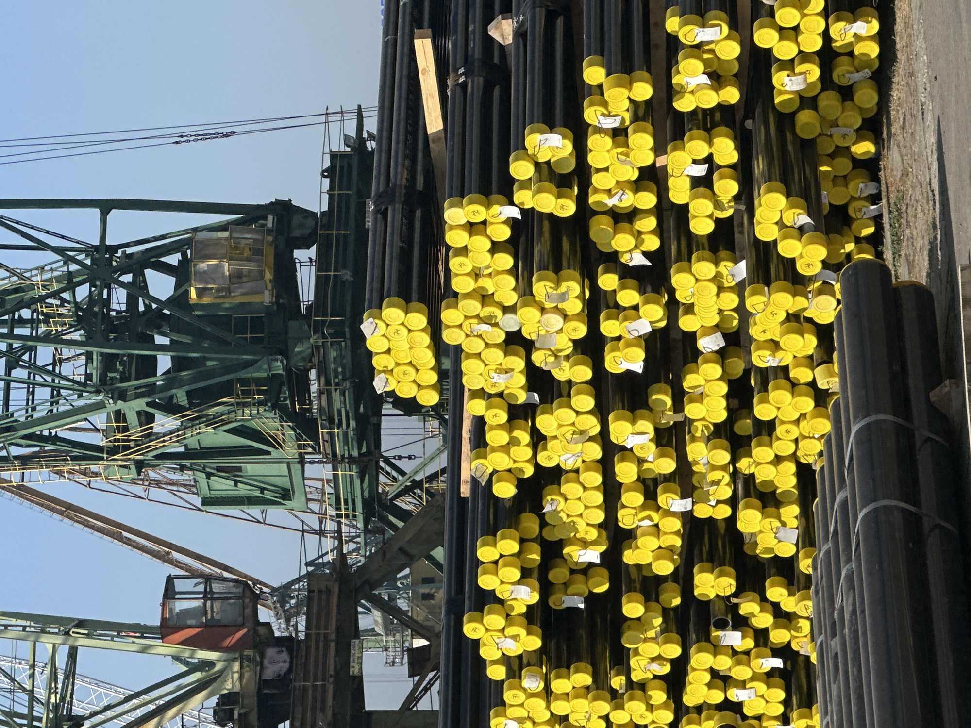 The image captures a bright, sunny day at a port or industrial yard. The foreground is dominated by stacks of black pipes, many of which are capped with vibrant yellow protective covers. These pipes are neatly organized, suggesting a storage or staging area. In the background, massive green industrial cranes loom against a clear blue sky. These cranes are complex structures with visible beams, platforms, and cabins, indicating heavy-duty machinery used for lifting and moving large loads. The overall scene conveys a sense of industrial activity and logistics.