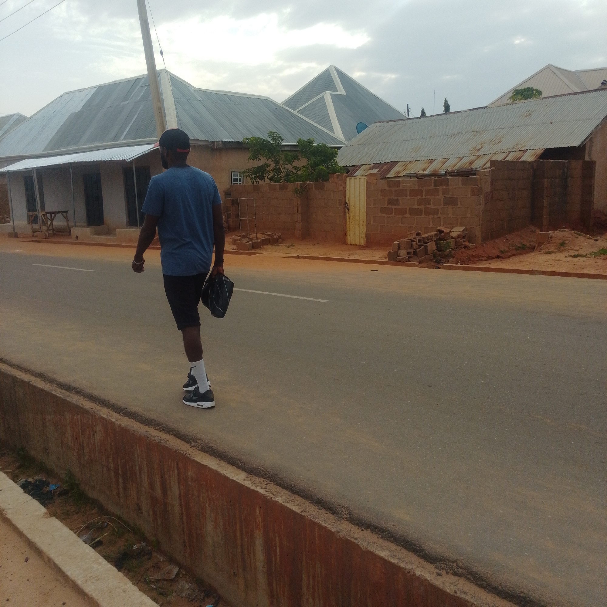 The image depicts a man walking away from the camera on a paved road in the city of Gombe, Nigeria. The man is wearing a blue t-shirt, black shorts, white socks, and black athletic shoes with white soles. He is carrying a black bag in his right hand. He is also wearing a dark baseball cap. The road has a single white line marking the center. To the left of the road is a concrete drainage ditch with some debris and vegetation. Beyond the ditch and road, there are buildings with corrugated metal roofs and brick walls. Some of the buildings appear to be residential or commercial structures. The sky is overcast with clouds. The overall scene suggests a typical urban or semi-urban environment.