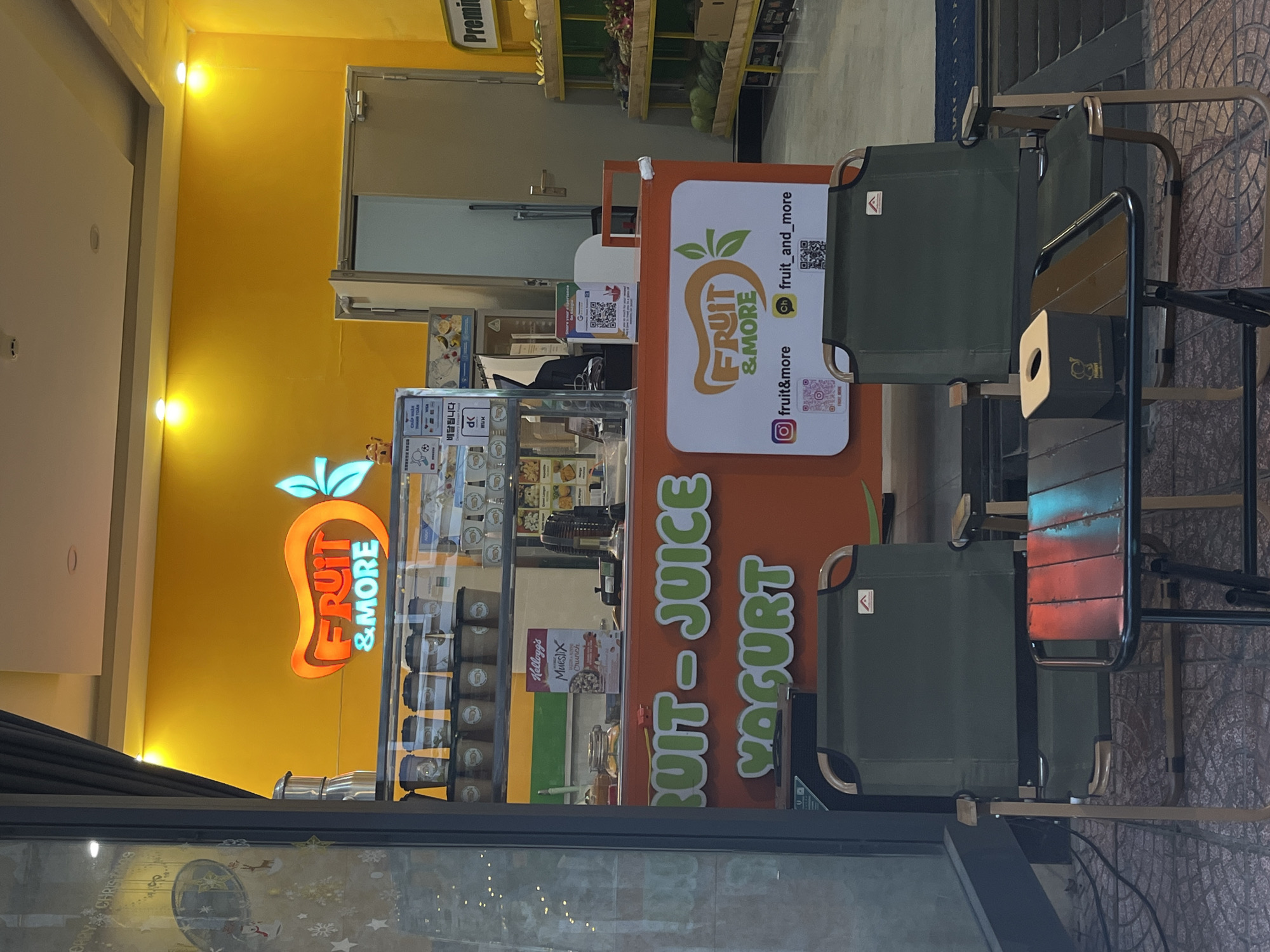 This image captures a vibrant, unoccupied "FRUIT & MORE" kiosk, likely situated in a commercial setting such as a mall or a shop front in Đà Nẵng, Vietnam. The scene features a brightly lit yellow wall with a prominent, glowing "FRUIT & MORE" neon sign shaped like a fruit on the left.
The central focus is an orange-colored kiosk, branded with the "FRUIT & MORE" logo and advertising "FRUIT - JUICE - YOGURT" in large green letters. Social media handles "@fruit&more" and "@fruit_and_more," accompanied by QR codes, are displayed on the kiosk's front, encouraging digital engagement. Within the kiosk's display, various containers, possibly holding fruit or yogurt products, and a "Kellogg's Mueslix Crunch" box are visible. A sign near the counter features Korean text "배달됩니다" (delivery available), indicating a service catering to Korean-speaking customers.
To the right, two dark green, foldable chairs with metal frames, accompanied by a small attached table holding a black tissue box, provide a simple seating area. In the background, shelving units are stocked with fresh produce like melons and other items, suggesting a larger fruit or grocery store section. A "Premi" (likely "Premium") sign is visible above these shelves. Reflections on a nearby glass surface show faint decorative elements, possibly holiday-themed. The entire scene is brightly illuminated, suggesting daytime or well-lit indoor conditions. No people are present, giving the impression of a moment before or after business hours, or a lull in activity.