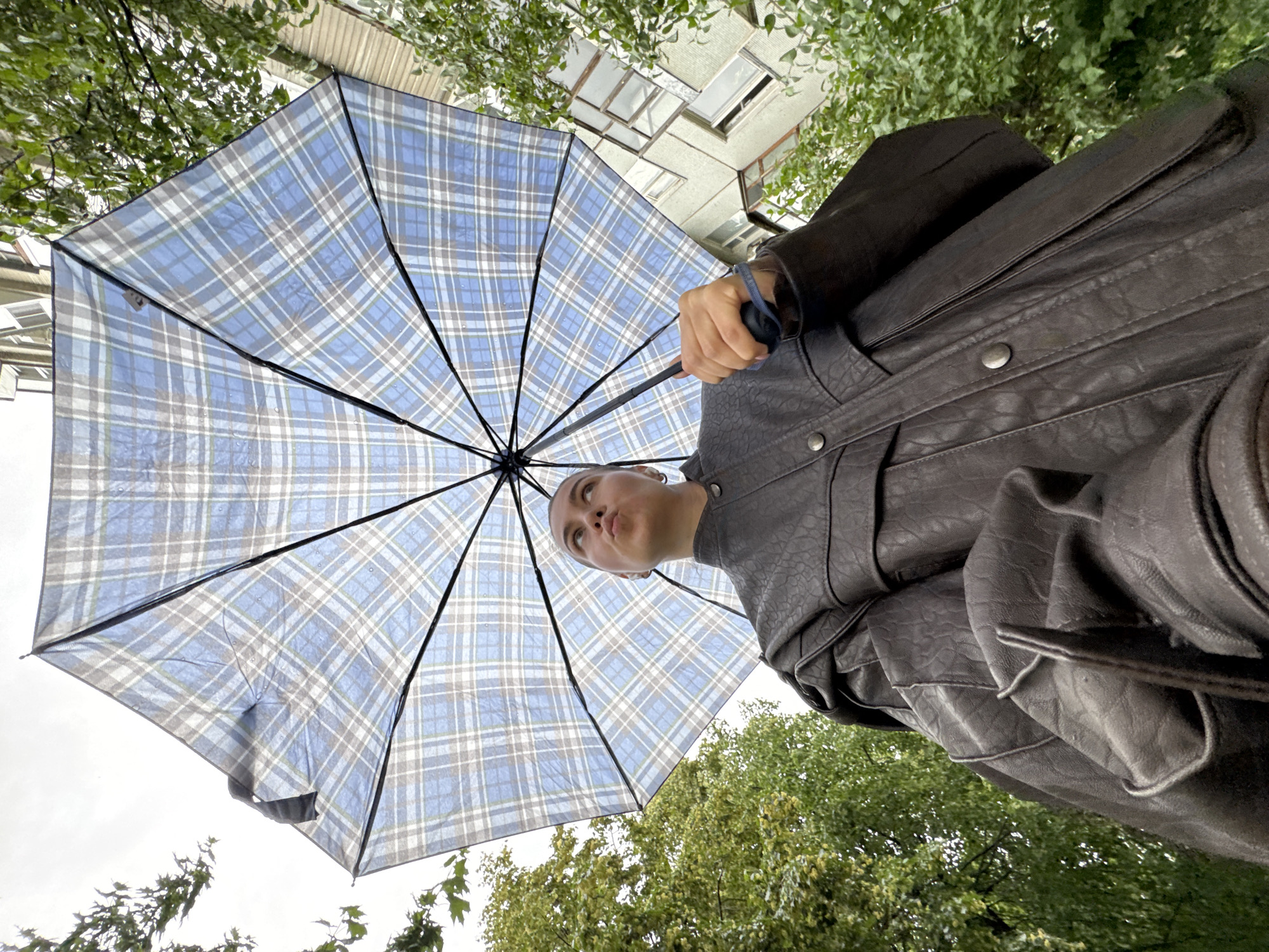 A low-angle, full-shot photograph shows a person holding an open, blue and white plaid umbrella over their head. The person is wearing a dark, textured leather jacket. They are looking up and away from the camera, with their lips pursed. The umbrella is positioned to fill the upper portion of the frame, obscuring a large part of the sky and the building behind the person. Green trees with lush foliage flank the right side of the image, and a section of a light-colored, multi-story building with numerous windows is visible to the upper right. The setting appears to be outdoors, possibly in an urban environment, on an overcast day.