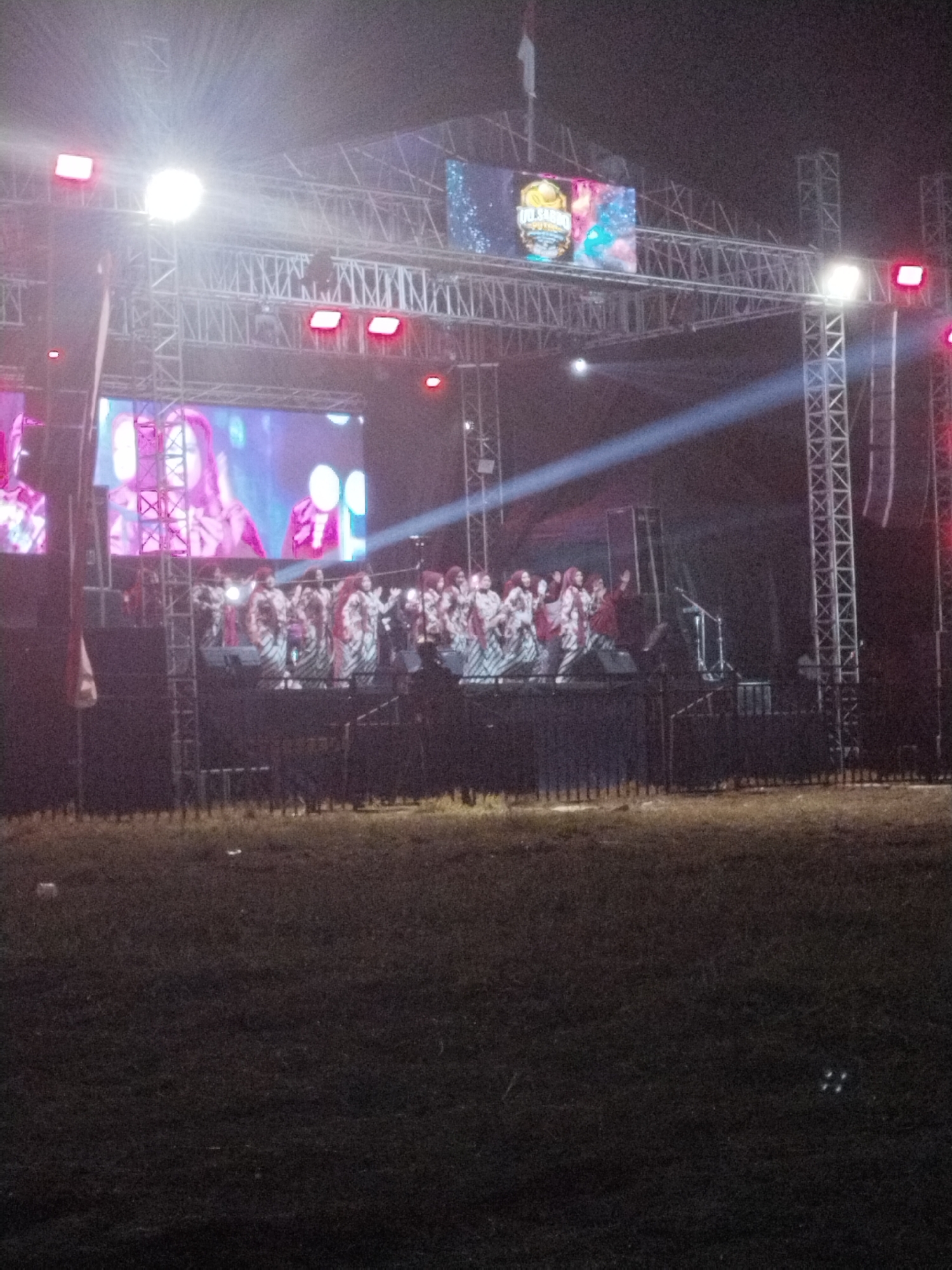 An outdoor stage is set up at night in Gringging, Indonesia. A group of performers, likely a choir or dance troupe, are on stage wearing matching outfits with red accents and patterned skirts. They are positioned in front of large screens displaying images of people. The stage is illuminated by bright lights, including spotlights that cast beams into the night sky. Truss structures support the lighting and screens, and speakers are visible. The area in front of the stage appears to be a field or open space with grass, and a fence or barrier separates it from the stage area. The overall scene depicts a performance or event taking place outdoors at night.