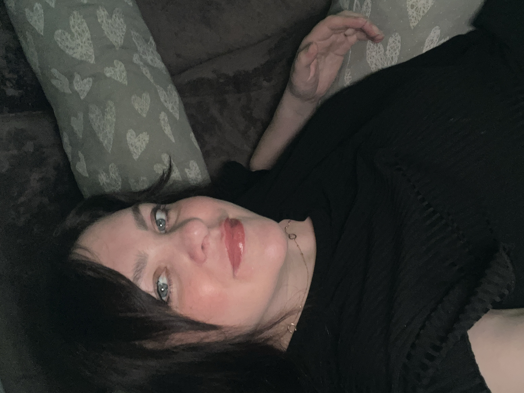 A woman with dark hair and fair skin lies on a brown couch with heart-patterned pillows. She wears a black top and a delicate gold necklace. Her eyes are light blue, and she has pink lipstick on. Her hand is raised slightly above her head. The environment is dimly lit. The location is not discernible based on the image.