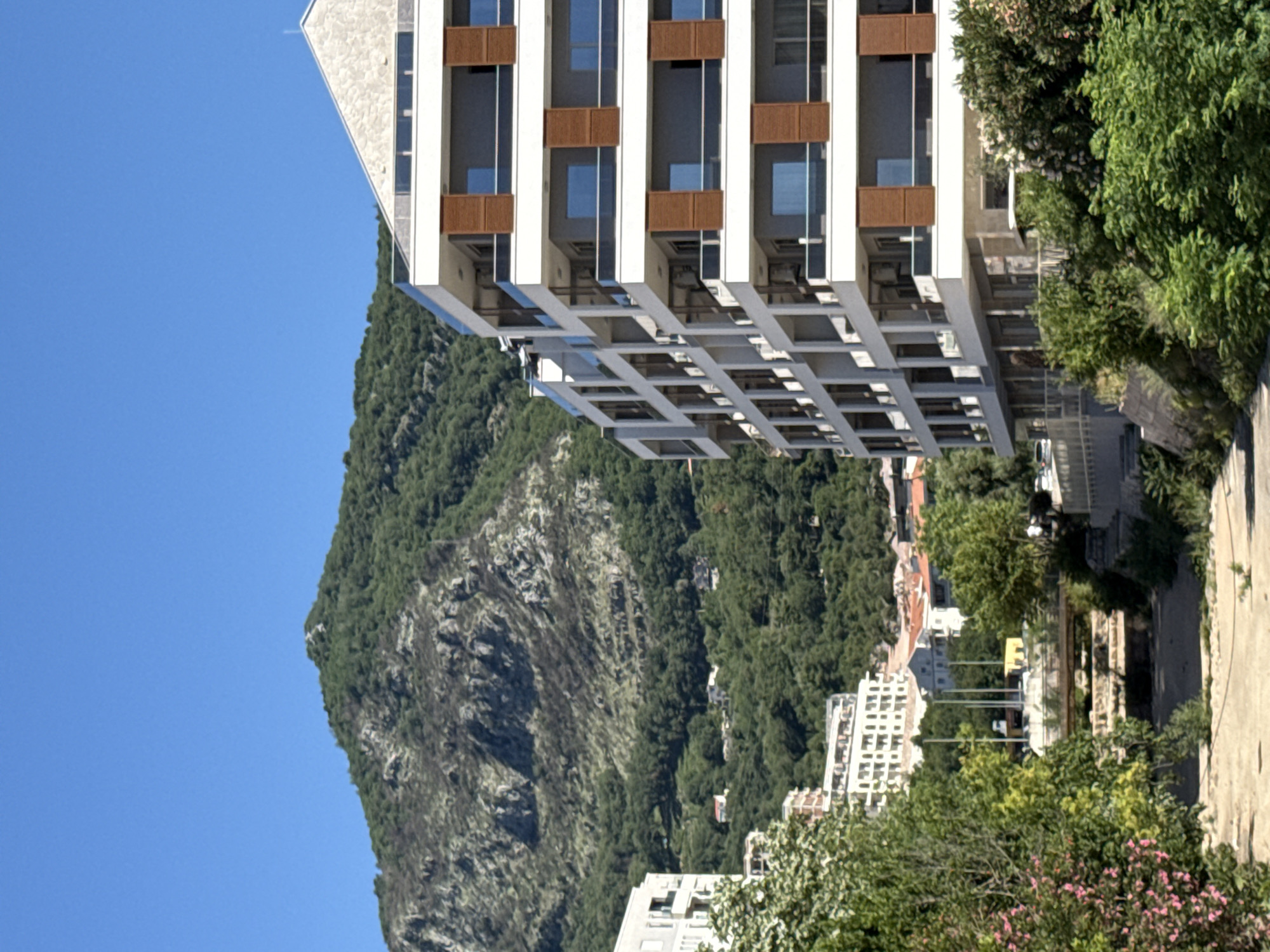 An architectural photograph depicts a high-rise building, likely residential, situated in the city of Budva, Montenegro. The building is characterized by its modern design, featuring multiple stories, recessed balconies, and a contrasting color palette of white and brown elements. The structure stands out against the natural backdrop of a prominent mountain partially covered in dense green vegetation, its rugged slopes etched with rock formations. A clear, bright blue sky fills the upper left quadrant of the image. The composition includes segments of other buildings, interspersed with trees and greenery, indicating an urban-meets-nature environment. In the foreground, there is some vegetation, and an elevated street to the right.