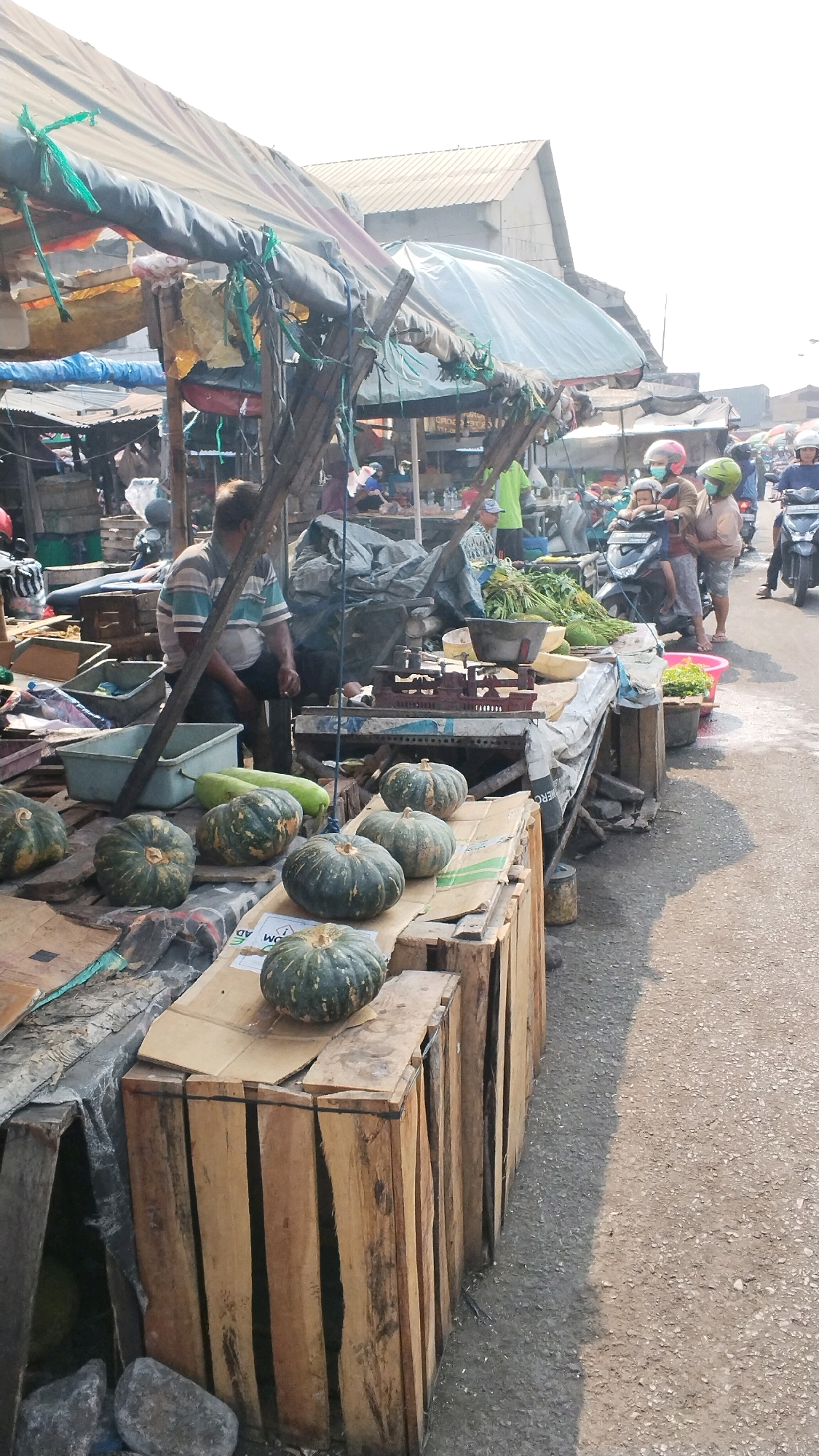 This is a street market scene in Mojokerto, Indonesia, under a bright, sunny sky. The market appears to be set up along a paved road, with several stalls selling produce. In the foreground, several green pumpkins are displayed on top of wooden crates. To the left of the pumpkins, a man in a striped shirt sits on a stool, looking away from the camera. Behind him, a blue plastic tub and other market supplies are visible.
Further into the market, more stalls are set up with awnings providing shade. Several people are present, some on motorbikes. One motorbike has a driver and a passenger with a child in the middle, all wearing helmets. Another person on a motorbike, also wearing a helmet, is seen in the distance. Various vegetables, including what appear to be long gourds and leafy greens, are displayed on tables. A scale is also visible on one of the market tables. The overall impression is of a bustling local market on a typical day.