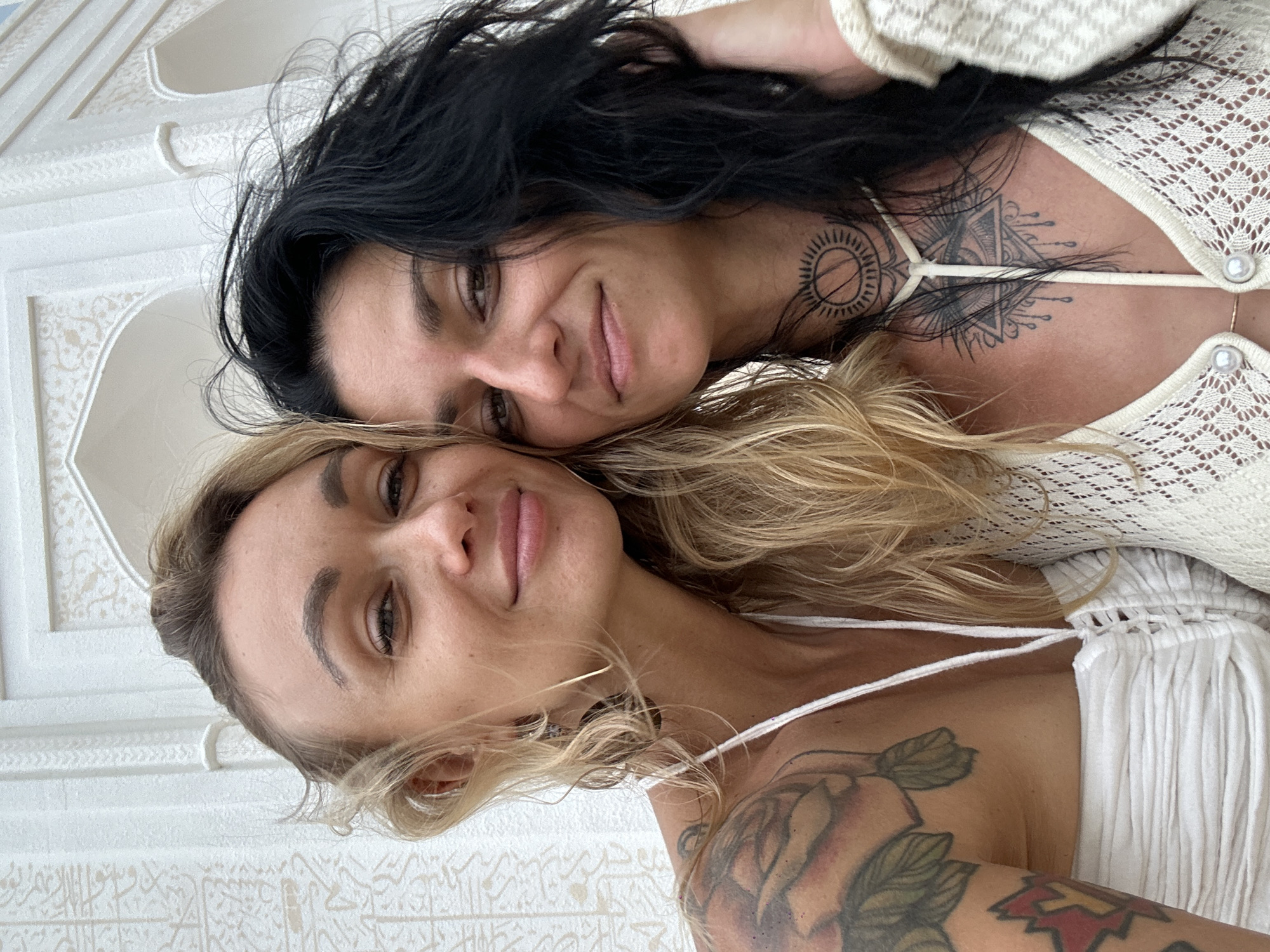 Two women pose closely together. The woman on the right has dark hair and fair skin, with tattoos visible on her neck and chest. She wears a white, crocheted top. The woman on the left has blonde hair, tan skin, and a tattoo on her left arm and chest. She wears a white top. They are indoors, with patterned walls and ceiling. The location is Mui Ne, Vietnam.