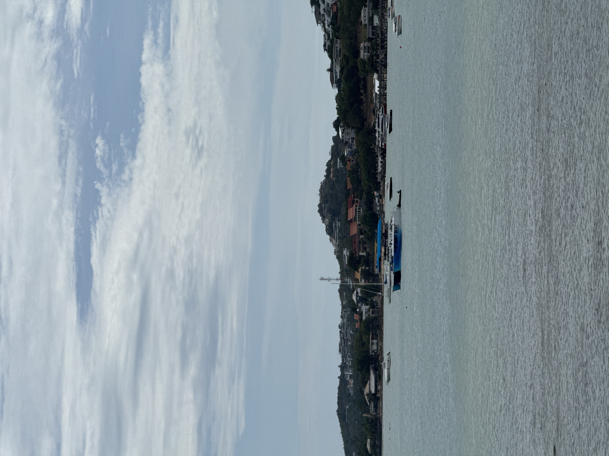 A body of water is in the foreground, with a landmass in the background. The landmass is a hill covered in lush green trees, with houses and buildings dotting its slopes. The houses are of various colors and architectural styles, suggesting a residential area. There are several boats visible on the water, including a sailboat, a catamaran, and several smaller motorboats. The sky is cloudy, with patches of blue peeking through. The overall scene depicts a picturesque coastal landscape. The location is Ko Samui District, Thailand.