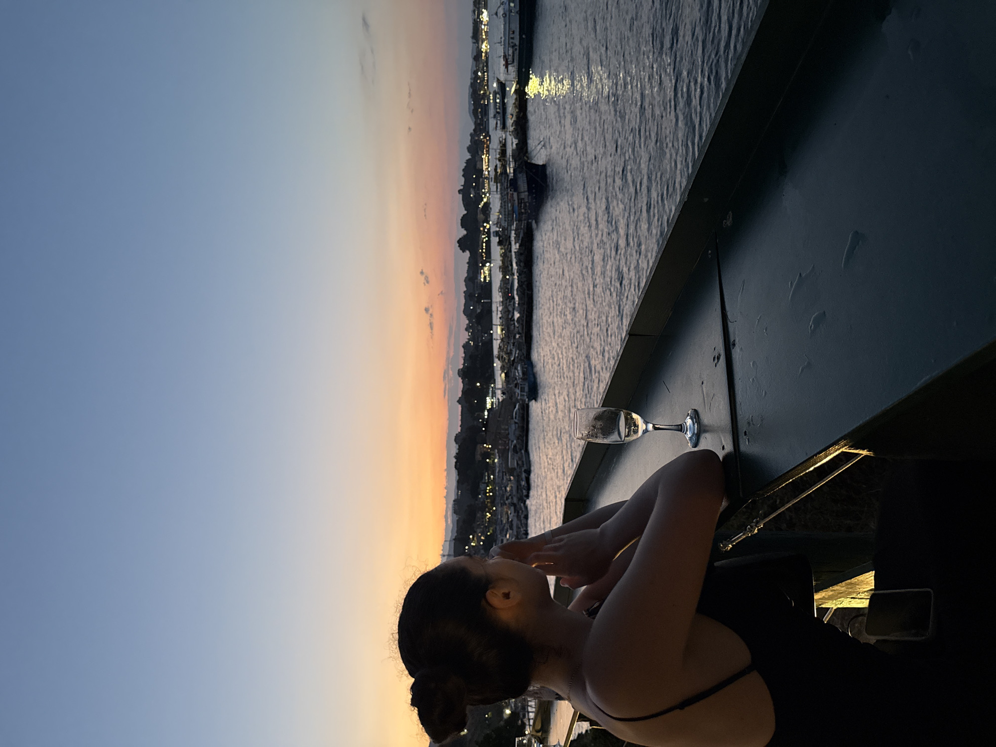 A woman with her hair in a bun, wearing a black dress, sits with her back to the camera, looking out at a harbor during sunset. She has her hands clasped together near her chin. A glass of white wine sits on a dark surface in front of her. The water of the harbor is calm, with several boats moored. The sky is a gradient of blue and orange, indicating sunset. Lights from a distant town twinkle along the coastline. The scene is peaceful and serene.