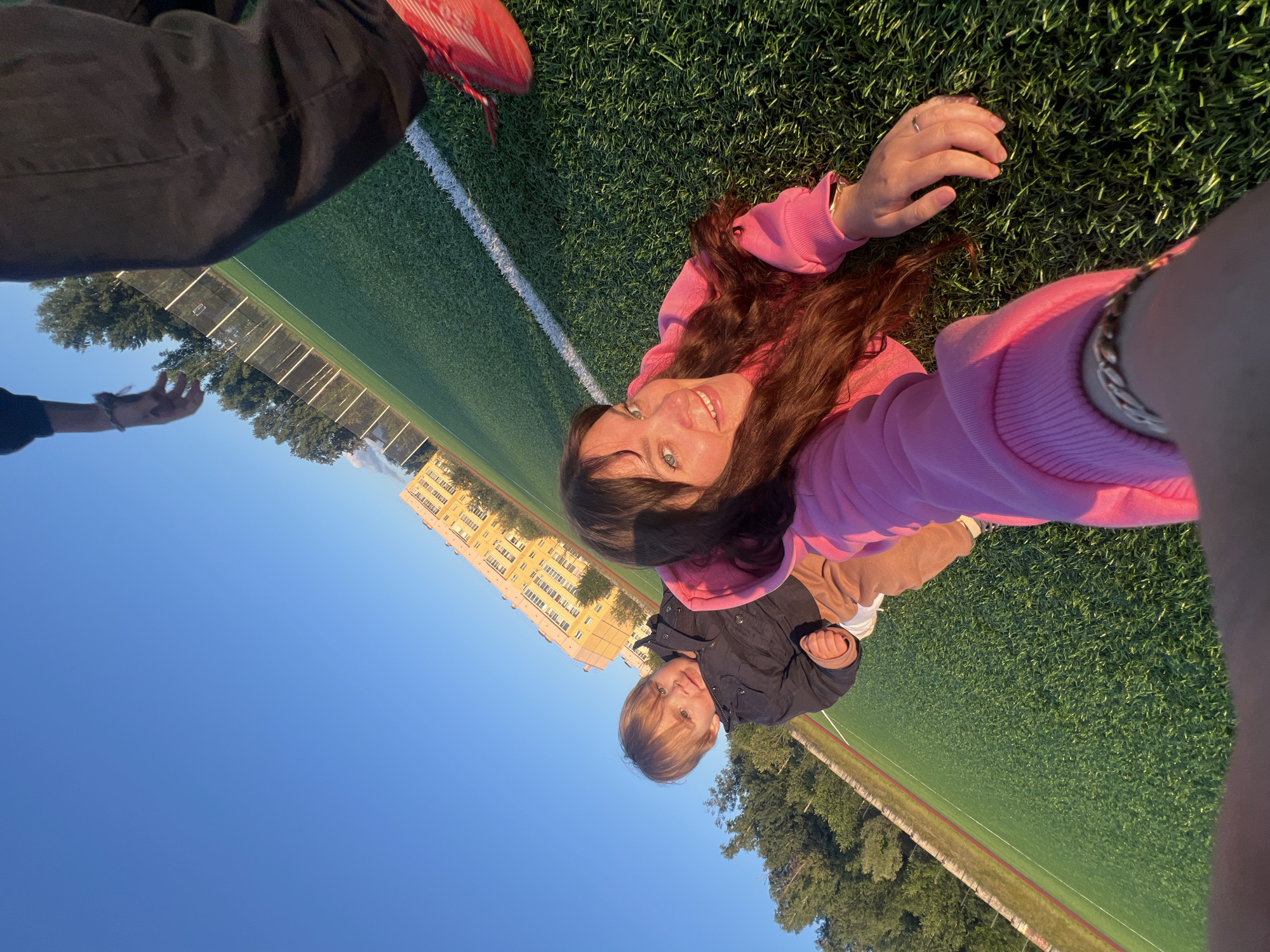 The image shows two people lying on a grassy field with a clear blue sky above. The person in the foreground is a woman with long brown hair, wearing a pink hoodie. She is smiling and looking at the camera, with her right hand resting on the grass. A man with blond hair, wearing a dark jacket, is lying next to her. A building with yellow walls and multiple windows is visible in the background, along with some trees and a fence. The sun appears to be low in the sky, suggesting it is either early morning or late afternoon, and the weather is clear and sunny. The setting appears to be a sports field, possibly a soccer field, given the presence of a white line marking the field. The overall mood seems relaxed and happy. There is no visible text in the image.
