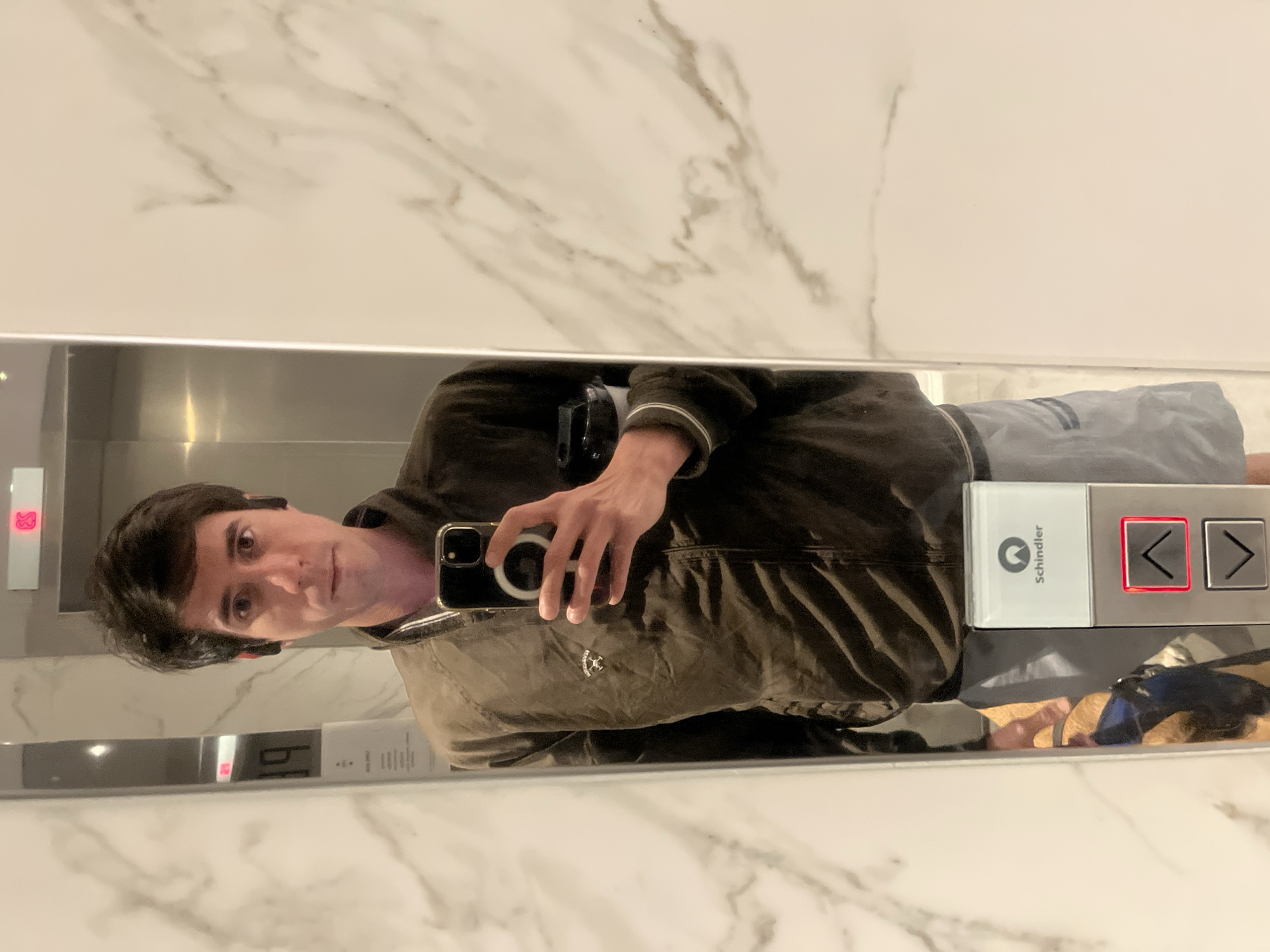 A man is taking a selfie in an elevator with marble walls, reflected in the mirror. He is wearing a brown jacket and gray shorts. The elevator has a Schindler control panel with an illuminated left arrow button. The location appears to be in the city of Buenos Aires, Argentina based on previous interactions.