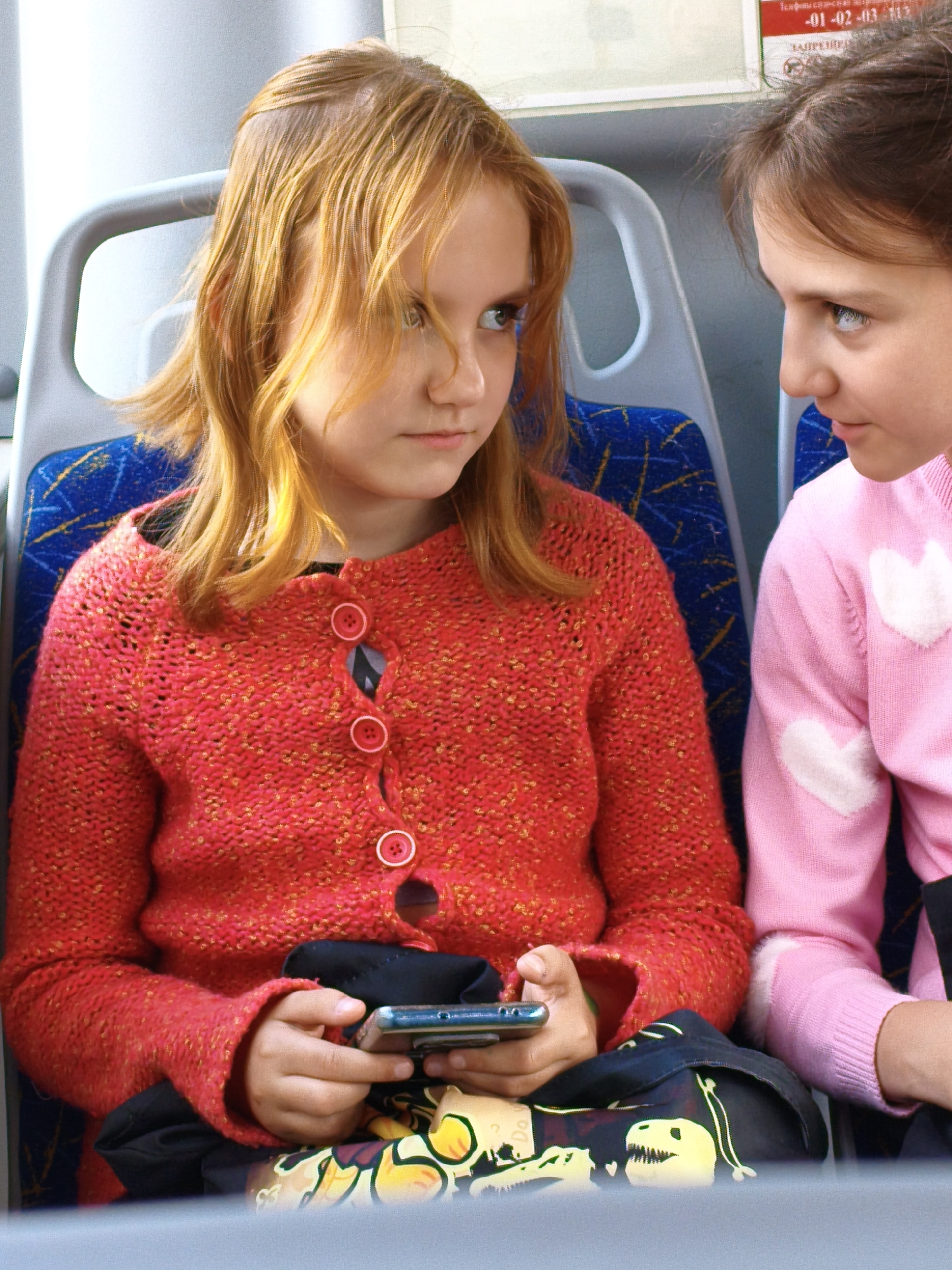Two young girls are seated side-by-side on a public transportation vehicle, likely a bus, in Orenburg, Russia. The girl on the left, with reddish-blonde hair, wears a red knit sweater with white buttons and is holding a smartphone in her hands. The girl on the right, with brown hair, wears a pink sweater with white hearts on it. Both girls are looking in different directions, suggesting they are either engaged in their own activities or observing their surroundings. The seats are blue with a yellow pattern, typical of public transport in Russia. The overall lighting suggests an indoor setting, possibly during daylight hours.