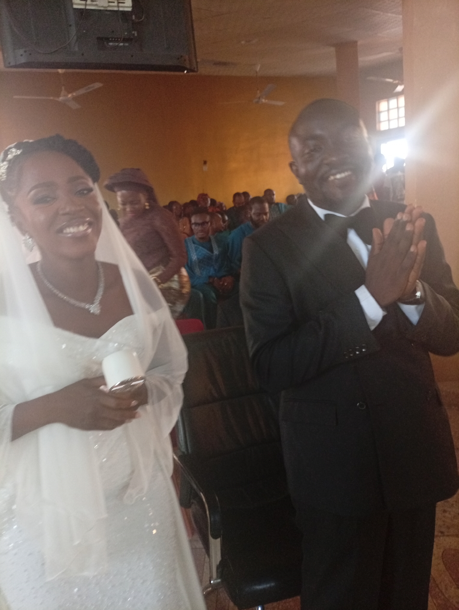 The scene is an indoor event in Sabon Tasha, Nigeria. Two primary subjects are positioned in the foreground, facing the camera. The subject on the left is a woman with dark skin, styled dark hair adorned with a sparkling hairpiece, wearing a white, sleeveless wedding gown featuring a sequined bodice and skirt. A sheer white veil extends over her shoulders and down her back. She wears a silver necklace with a central pendant and matching earrings. Her facial expression shows a broad smile. In her left hand, she holds a small, cylindrical white object with a metallic base. The subject on the right is a man with dark skin, wearing a black suit, white dress shirt, and a black bow tie. His hands are clasped together at chest level, and he wears a silver watch on his left wrist. His facial expression also shows a broad smile.
In the background, several individuals are visible, seated in dark chairs. Behind the woman, a person wearing a brown hat is partially obscured. Further back, individuals are seen in various attire, including blue and patterned garments; one person on the right wears eyeglasses. The walls of the venue are a uniform light brown or golden color. Two white ceiling fans are visible, one in the upper left and another near the center-right. A large, dark flat-screen monitor is mounted high on the wall in the upper left. A bright light source, likely a window, is present on the far right, creating a significant glare. The flooring is not clearly discernible. The immediate foreground seating consists of dark, padded chairs.