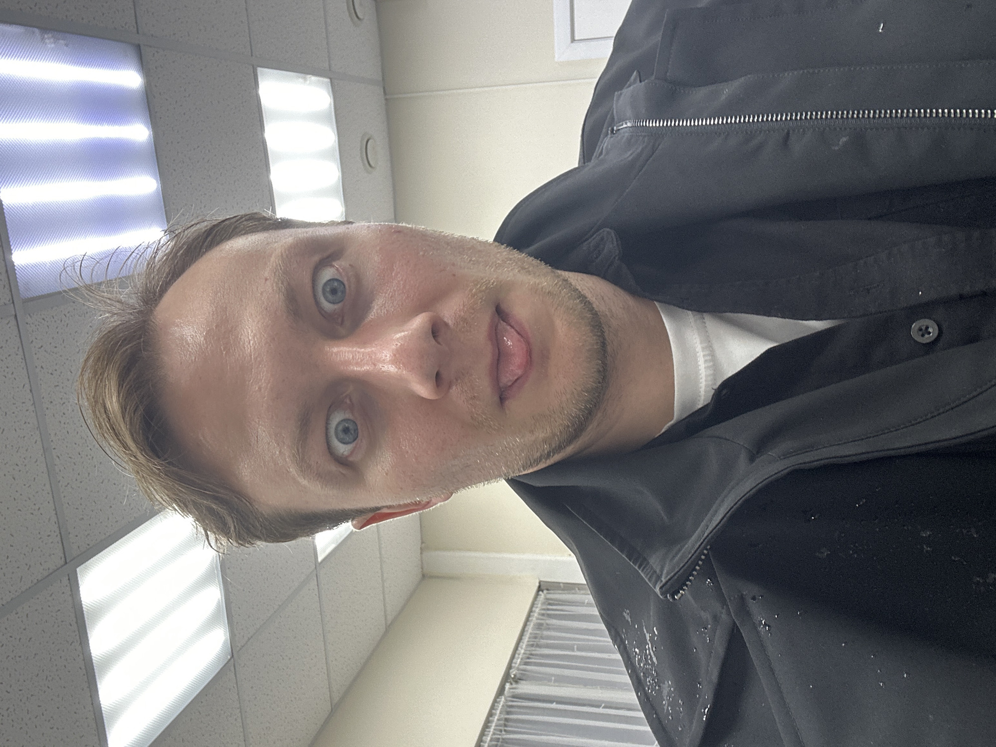 A close-up, upward-angled selfie of a Caucasian male with light brown hair and blue eyes. He is sticking his tongue out slightly. He is wearing a black jacket over a white shirt. The jacket appears to be wet.
The background consists of a tiled ceiling with fluorescent lights and a light-colored wall. Based on the environment's appearance, the location is most likely an office or commercial space. There is no information in the image to place the location in the city of Sochi, Russia.