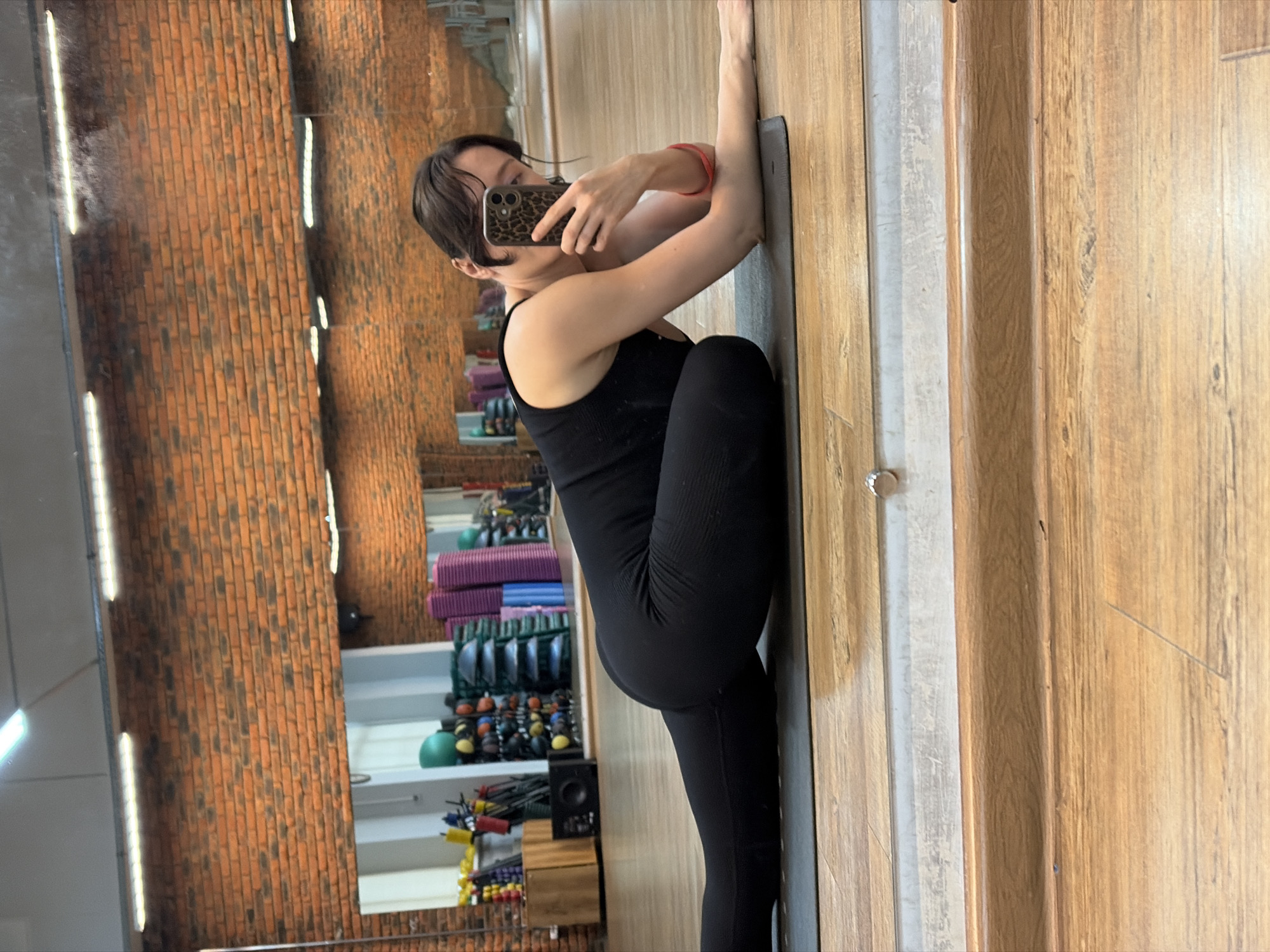 The image shows a young woman in a black workout outfit performing a yoga pose on a mat in a gym. She is leaning against a wooden wall and taking a selfie with her phone. The gym has a brick wall on one side and a mirror reflecting exercise equipment, including yoga mats and weights. The lighting suggests it is daytime. The woman appears focused on her pose. The location is identified as Chaikovsky, Russia.