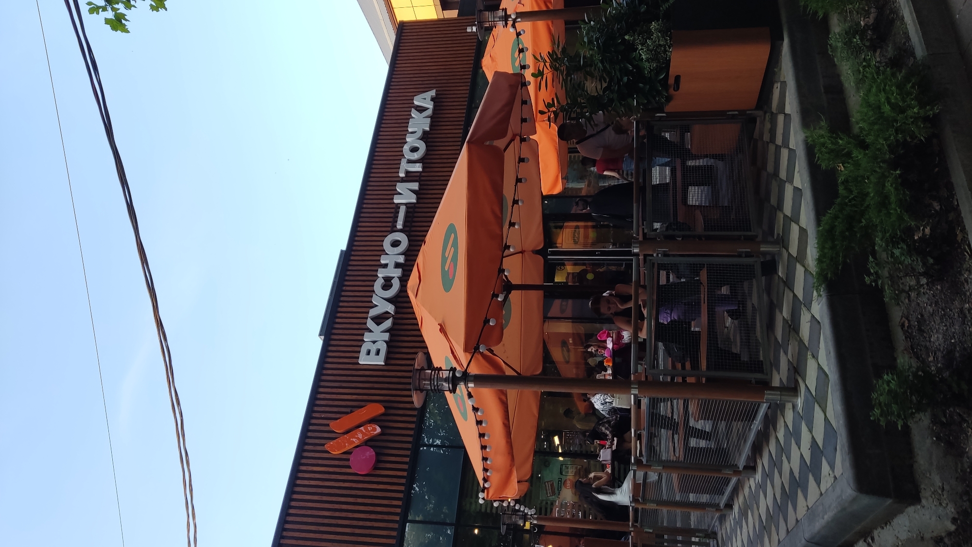 The image shows the exterior of a fast-food restaurant, "Vkusno - i Tochka," located in the city of Krasnodar, Russia. The restaurant features a modern design with a facade of vertical wooden slats and large glass windows. Prominently displayed on the facade is the restaurant's name in white lettering, along with its logo, which consists of stylized pink and orange shapes resembling food items.
In front of the restaurant, an outdoor seating area is visible, covered by large orange umbrellas adorned with the restaurant's logo. Several tables and benches are set up under the umbrellas, with some patrons seated and others visible through the windows. The flooring of the outdoor area is a checkered pattern of black and white tiles.
Several people are present in the scene, some dining outdoors and others walking by. The overall atmosphere appears to be casual and relaxed, with people engaging in everyday activities. The image is captured during daylight hours, with a clear blue sky overhead and some trees visible in the background. The lighting suggests it might be late afternoon or early evening.