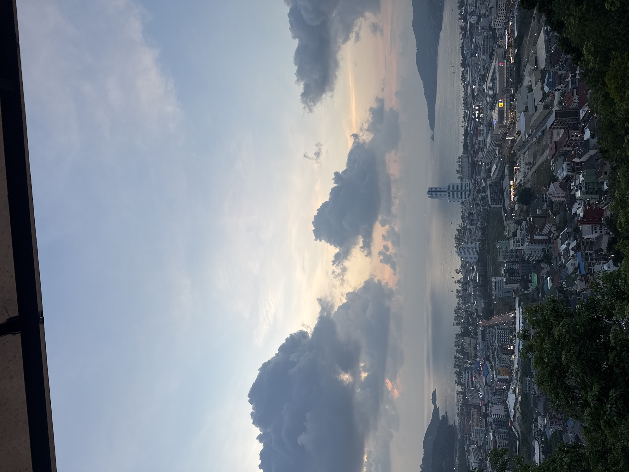 The image presents a panoramic aerial view of Patong, Thailand, captured at dusk from an elevated vantage point. The scene encompasses a dramatic sky, a sprawling coastal city, and the sea stretching to the horizon.
The sky occupies a significant portion of the frame, displaying a gradient of colors from light blue overhead to warmer, soft orange and golden hues near the horizon, indicative of either sunset or sunrise. Large, voluminous dark grey clouds are prominent, scattered amidst lighter, more diffused clouds, creating a dynamic and moody atmosphere.
Below the sky, the calm waters of what appears to be the Andaman Sea extend across the mid-ground. Distant mountainous landmasses or islands are visible on the far side of the bay, silhouetted against the lighter horizon.
The foreground and mid-ground feature the dense cityscape of Patong, with numerous buildings of varying heights and styles lining the coastline. Many city lights are already illuminated, signaling the transition into evening. A distinctive tall, white high-rise building stands out near the water's edge within the urban sprawl. On the far right, dark green foliage, likely from trees or bushes, partially frames the view, suggesting the photograph was taken from a hillside or a high-rise balcony. A dark, broad architectural element, possibly a roof overhang or balcony railing, frames the upper-left corner of the image.
The overall impression is one of a vibrant coastal city winding down as evening approaches, set against a backdrop of a picturesque, cloudy sky. No specific activities or visible text are discernible in the image.