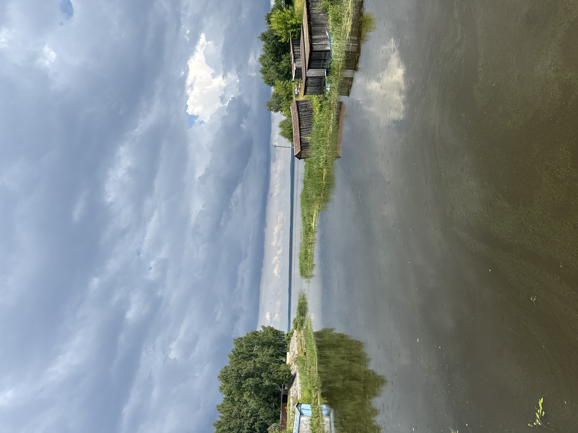 The image depicts a waterfront scene in Chastyye, Russia, characterized by a body of water reflecting a cloudy sky. On the left bank, there are lush green trees bordering a stone embankment, with a small blue structure and another lighter colored building partially visible. The water itself is dark and calm, with noticeable ripples indicating light rain. Scattered aquatic vegetation is present in the water. On the right bank, several rustic wooden sheds with corrugated metal roofs are lined up along the grassy shore, some appearing to be boat houses. The sky is filled with dramatic, dark grey storm clouds interspersed with patches of lighter sky and some white clouds. The overall impression is a tranquil yet somewhat brooding landscape under an overcast sky.