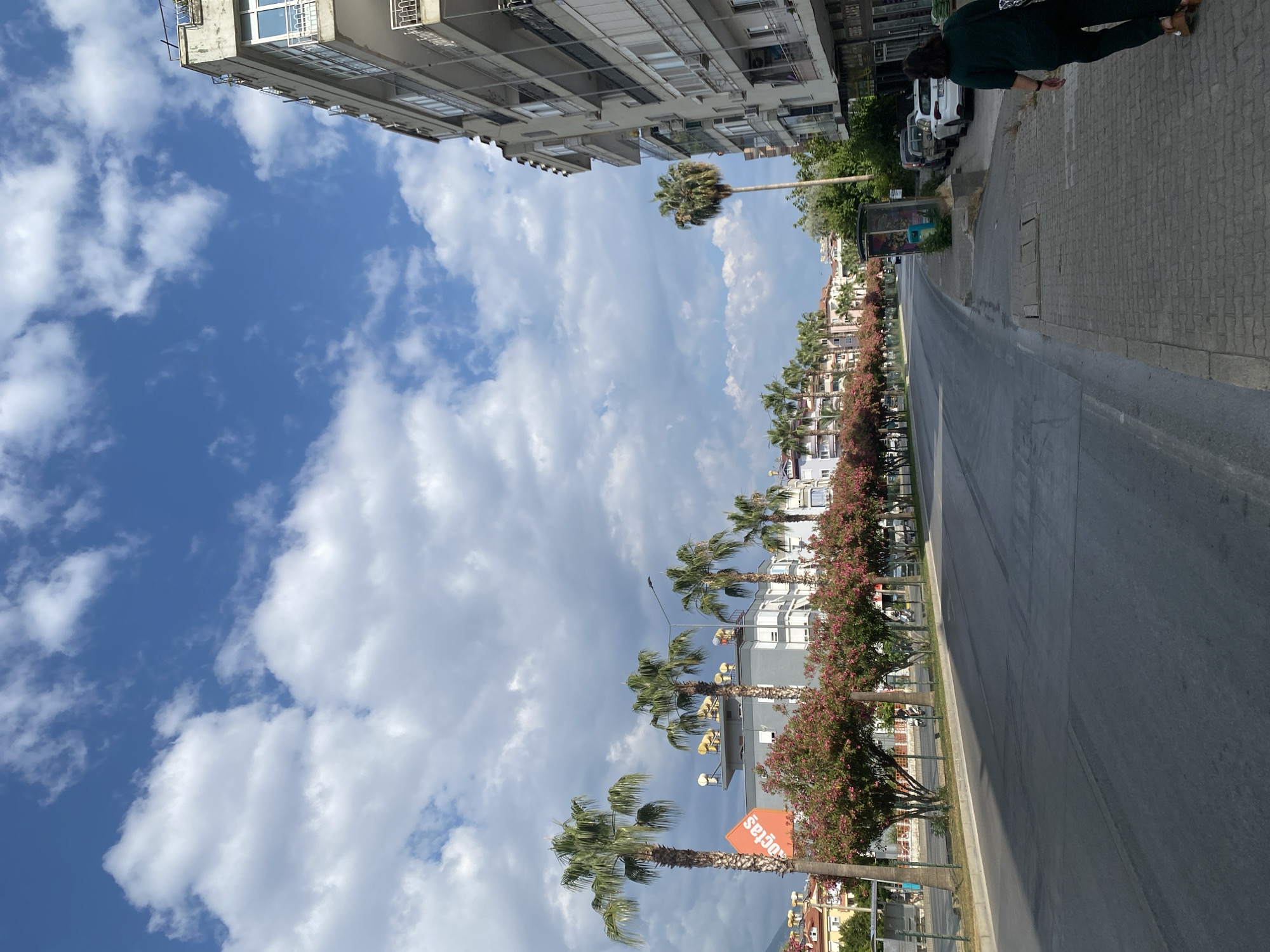 The image captures a street scene in Alanya, Turkey, likely during the daytime given the bright sky and shadows. The perspective is from a street-level viewpoint, looking down a road lined with buildings and palm trees.
On the right side of the road, a multi-story building dominates the foreground, exhibiting a modern architectural style with balconies and windows. A person, dressed in dark green clothing, is walking away from the camera along the sidewalk. Several parked cars are visible along the curb in front of the building.
To the left of the road, a row of buildings with shops on the ground floor are visible. Palm trees and flowering trees with pink blossoms line the sidewalk and street, creating a tropical or Mediterranean atmosphere. One prominent building on the left features an orange advertisement with white text, possibly a business logo. Solar water heaters are visible on the rooftops of some buildings.
The road itself appears to be asphalt and shows some wear and tear, with visible tire marks. The sky is a bright blue with scattered white clouds, suggesting fair weather. In the background, more buildings and some hills or mountains are visible, characteristic of Alanya's landscape. The presence of a bus stop shelter further indicates an urban or semi-urban setting. The overall impression is a bustling street in a warm climate.
