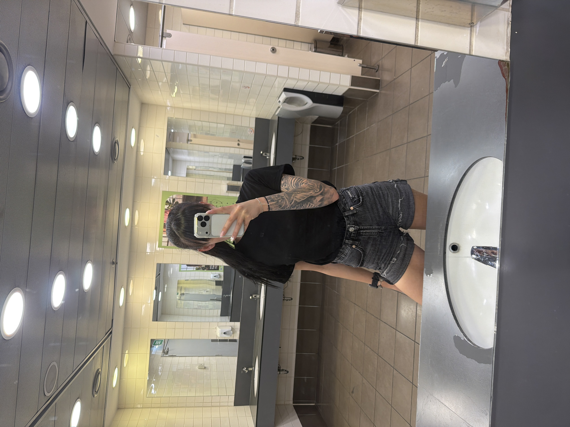 The image shows a person taking a selfie in a public restroom. The person is wearing a black t-shirt and denim shorts, with tattoos visible on their arm. They are holding a smartphone to take the picture. The restroom appears to be modern, with white tiled walls, grey countertops, and multiple sinks. There are also urinals and stalls visible in the background. The lighting is bright, coming from overhead circular fixtures. The floor is tiled with a beige and brown pattern. The image does not contain any information that would allow for its location to be identified as Plovdiv, Bulgaria.