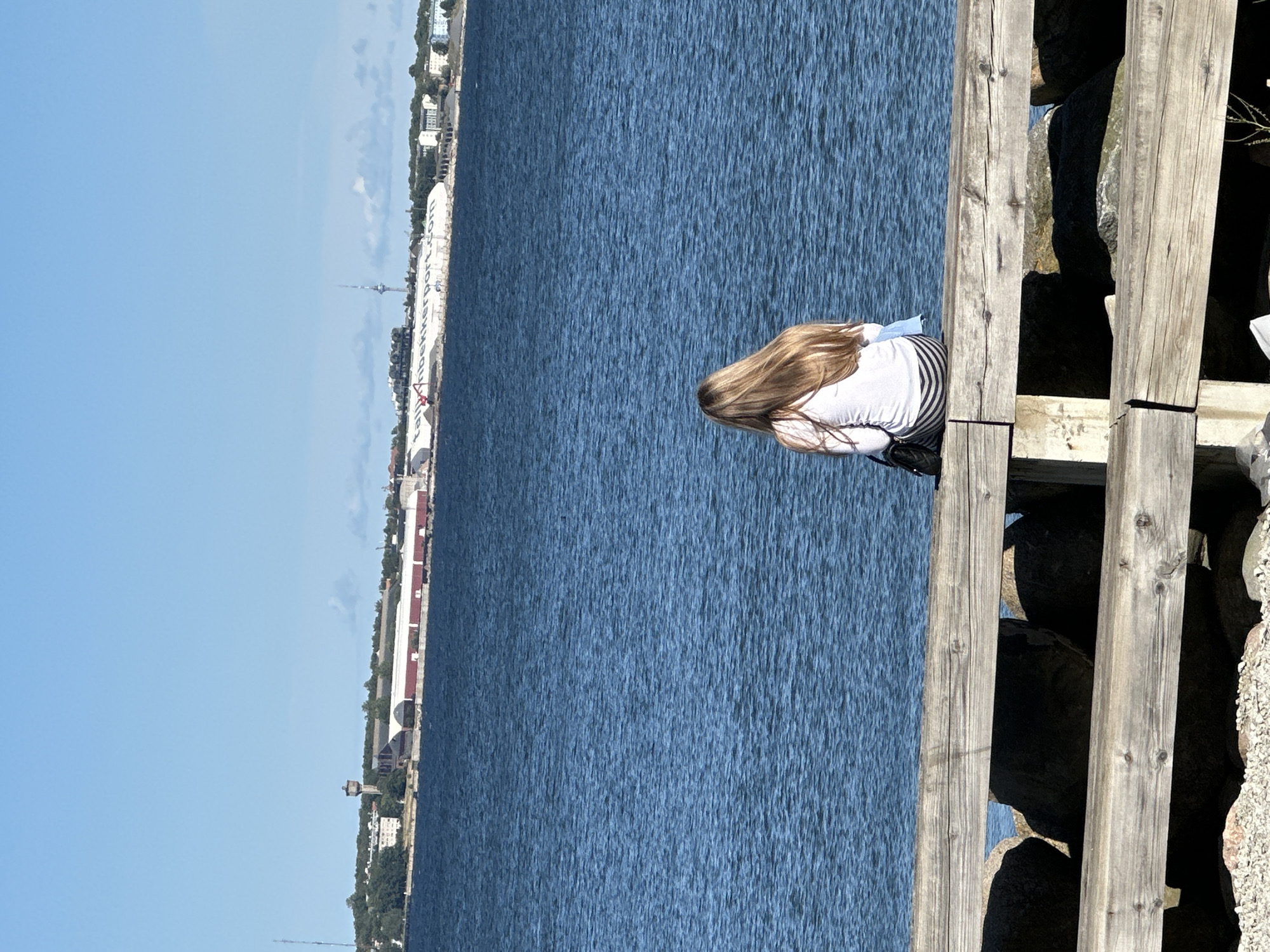 The image shows a young woman with long blonde hair sitting on a wooden pier, facing the water. She is wearing a white long-sleeved shirt and black and white striped pants. A black purse is visible beside her. In the background, across a body of blue water, is a cityscape with various buildings, including a tall antenna, a water tower, and industrial structures. The cityscape appears to be the Port of Tallinn, Estonia, as suggested by the presence of industrial port facilities and infrastructure. The sky is clear and blue. The foreground of the image is dominated by the wooden pier and large rocks.
