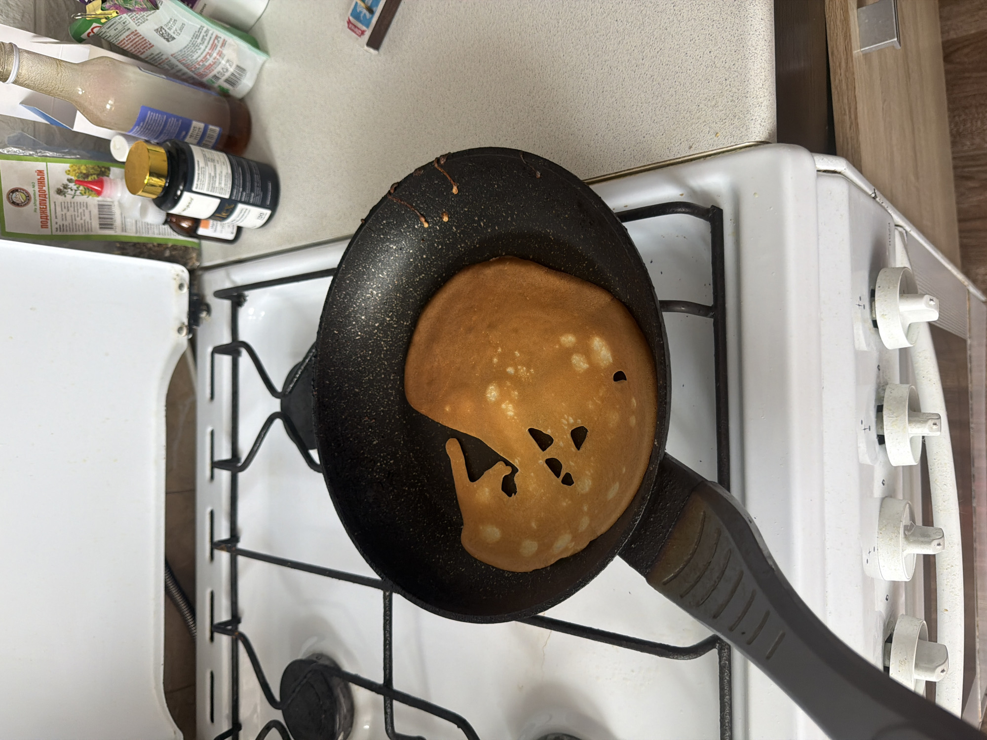 The image shows a pancake being cooked in a black frying pan on a white gas stove. There are several holes in the pancake. The stovetop has a black grate. The background includes various bottles and containers, along with the kitchen countertop and adjacent wall. The image was taken indoors, likely in a residential kitchen. There is no information about the location in the city of Leningradskaya, Russia.