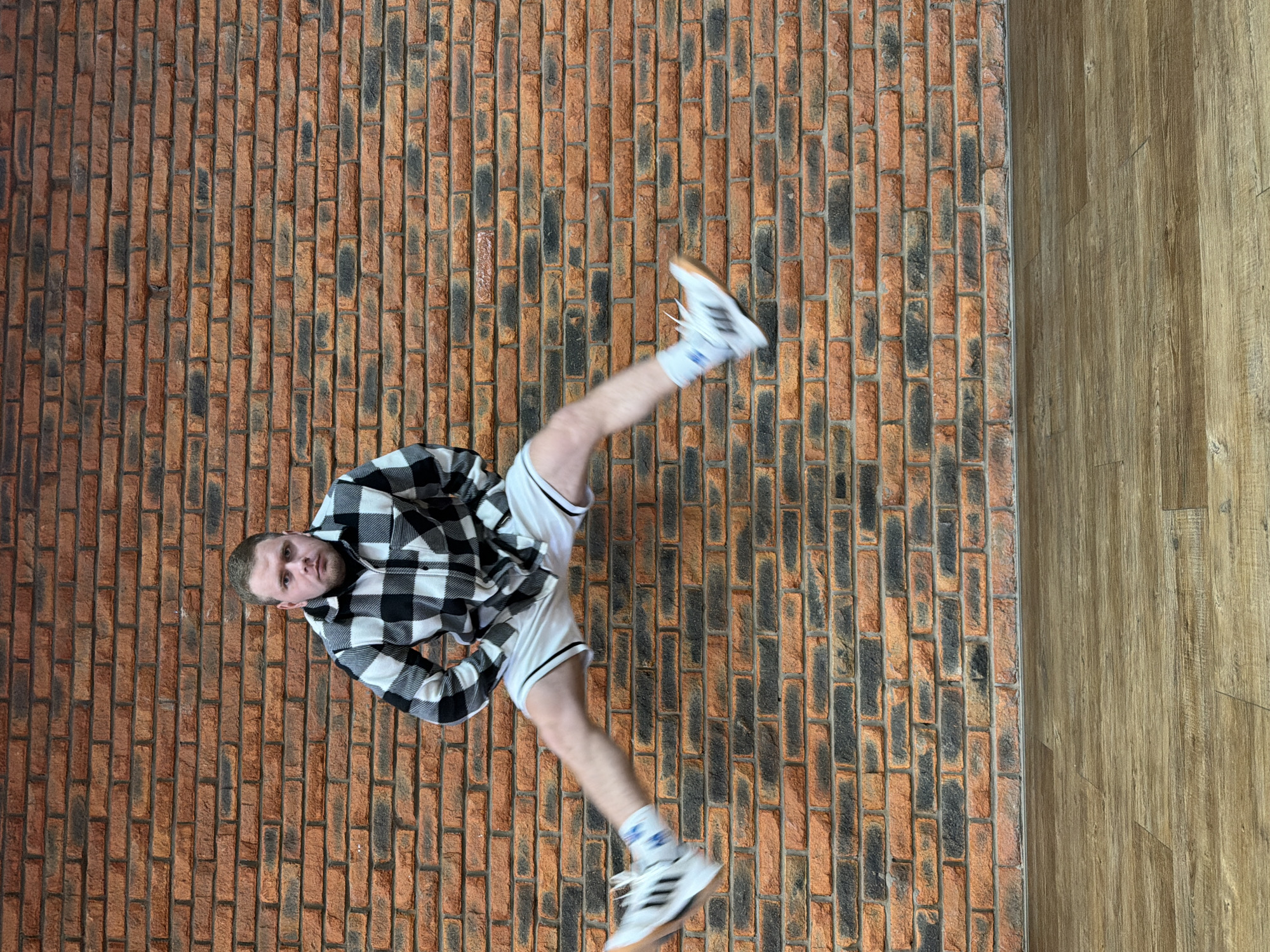 An indoor, vertically oriented shot captures a young man performing a dynamic pose, as if kicking upwards or falling, against a patterned background. The image is rotated 90 degrees clockwise from what might be its original orientation, presenting the man diagonally across the frame.
The subject is a light-skinned man with short, light brown hair, looking directly at the camera with a neutral to slightly serious expression. He is wearing a black and white plaid long-sleeved button-up shirt, white athletic shorts with black trim along the sides, white socks, and white athletic shoes with a tan/gum sole. The shoes clearly display the Adidas brand logo with three stripes on the side. Blue text or a logo is also visible on his white socks.
The background consists of two distinct sections. Approximately two-thirds of the left side is a textured surface resembling a brick wall or floor, with individual bricks in shades of reddish-brown and darker, almost black, patches, separated by lighter mortar lines. The right third of the background is a lighter brown wooden surface, possibly a floor or wall, showing visible grain and plank lines. The boundary between the brick and wood is a clear, straight line. No other objects or text are discernible in the background. The lighting suggests an indoor setting, likely with artificial or diffused natural light. The scene depicts a dynamic moment, possibly from a dance, martial arts, or gymnastics routine, set against a contrasting architectural backdrop in Chaikovsky, Russia.