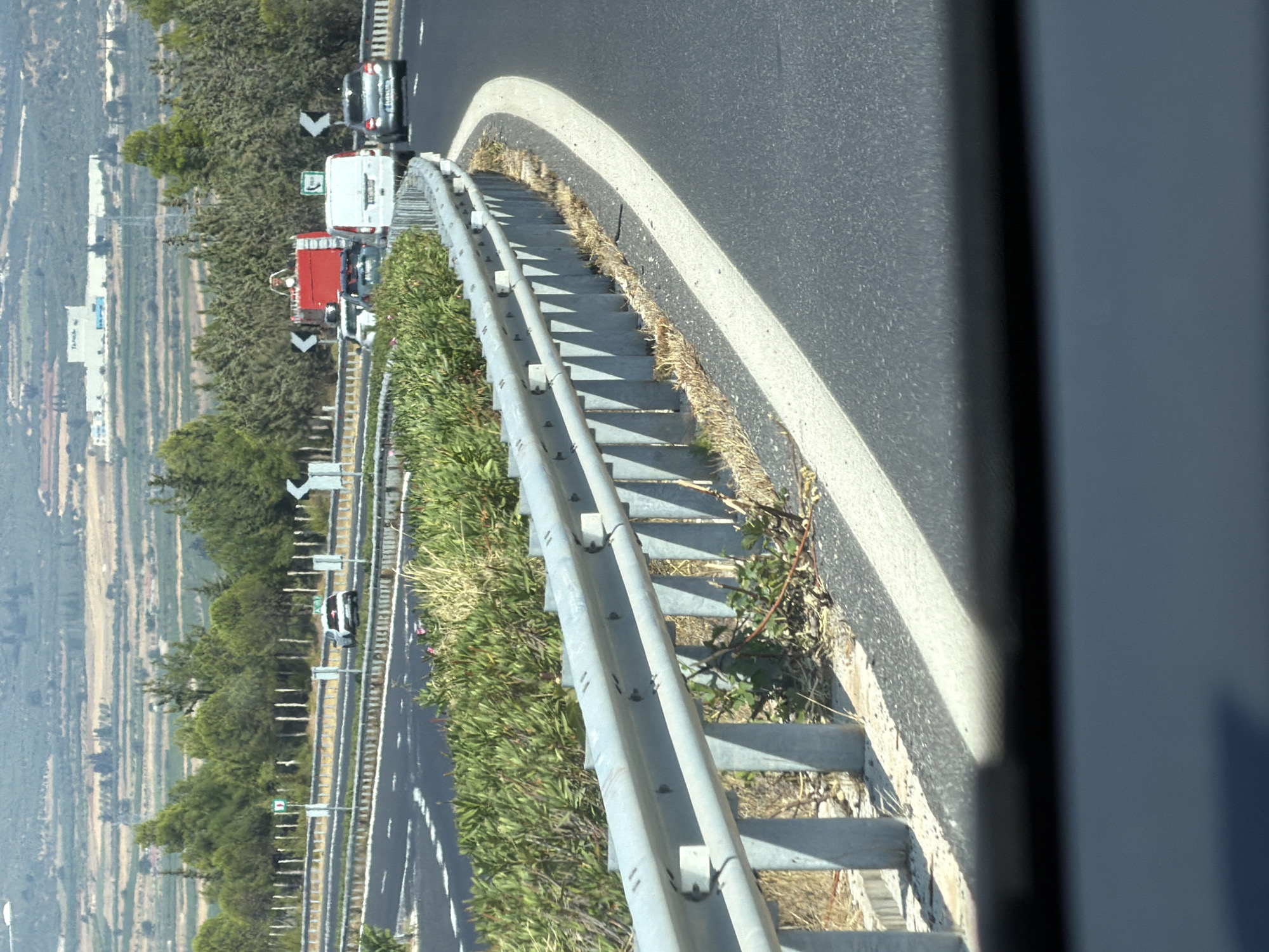 The image shows a highway in the Municipality of Nemea, Greece, with several vehicles on the road. In the foreground, a grey metal guardrail lines the side of the highway, with a white stripe painted on the asphalt next to it. Beyond the guardrail, there is dense green vegetation. Further down the highway, a red fire truck with a ladder is visible, along with a white van and a silver car. In the background, there are hills with agricultural fields and a large building. The sky is overcast. The time of day appears to be daytime.