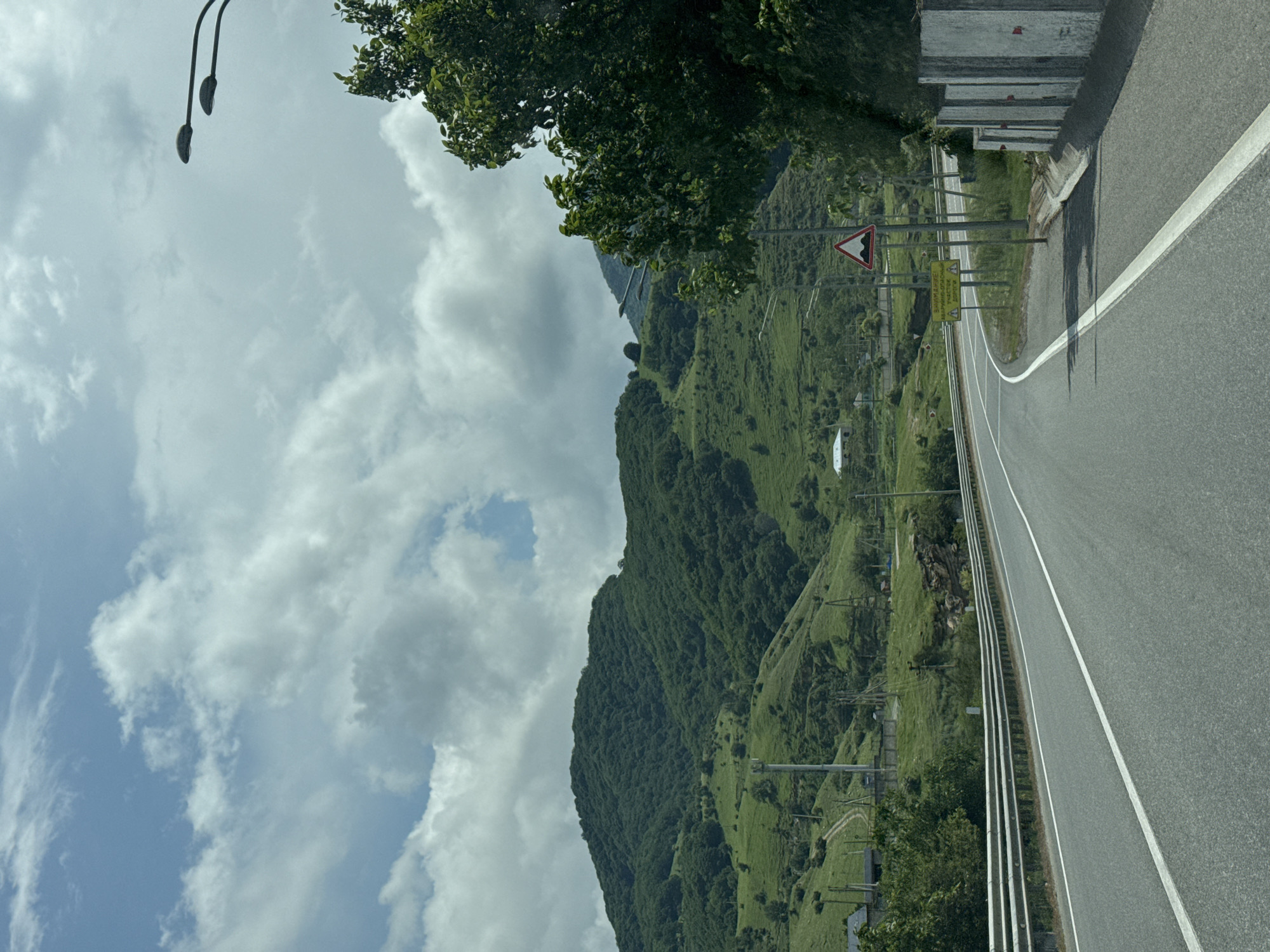 A highway curves through a verdant, hilly landscape under a partly cloudy sky. The road is paved with asphalt and marked with white lines. On the left, a metal guardrail runs alongside the highway. The hills are covered in lush green grass and trees, with some evidence of human settlements and structures, including power poles and what appears to be a small building. In the distance, the rolling hills rise to form larger mountains. On the right side of the road, a large tree with dark green leaves partially obscures the view of the sky and distant mountains. Several streetlights are visible along the highway. A triangular traffic sign depicting speed bumps is also present, along with a yellow sign with text that is difficult to read. Concrete barriers line the right edge of the road. The scene is reminiscent of a rural or mountainous region within the city of Ordzhonikidzevsky, Russia, given the terrain and vegetation. There are no people or animals visible in the image.