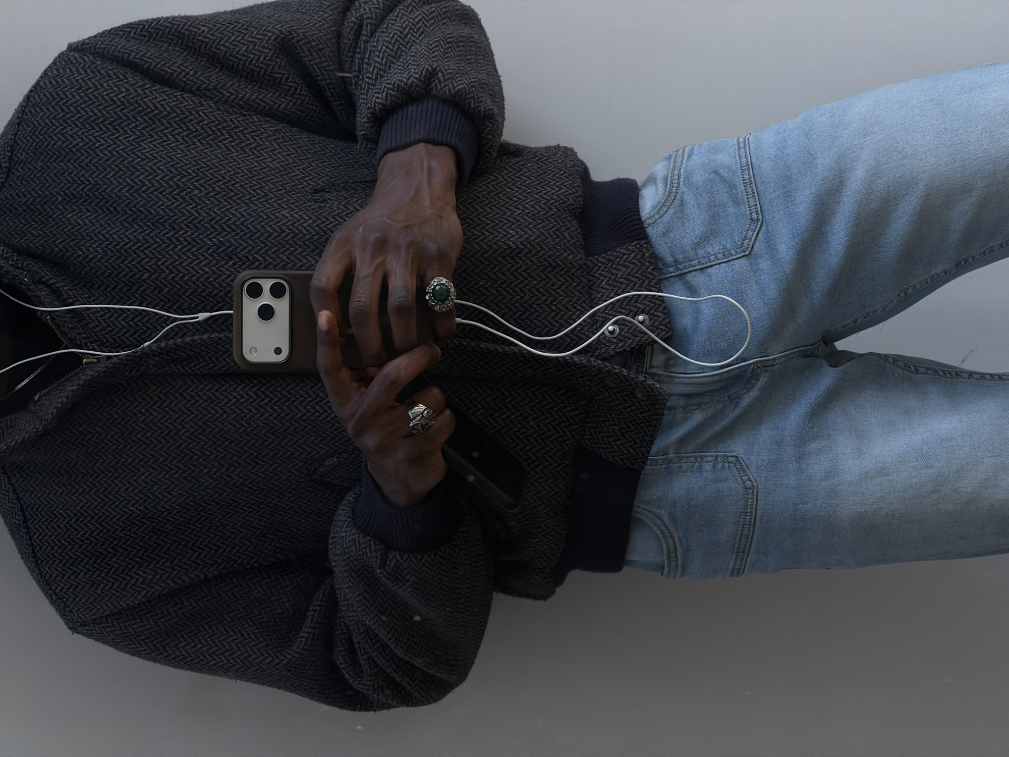 The image shows a person wearing a dark grey jacket with a herringbone pattern, light blue jeans, and two rings. The person's hands are holding a phone with white earbuds connected to it. The background is a plain grey surface. The scene appears to be indoors. The location is not discernible from the image.