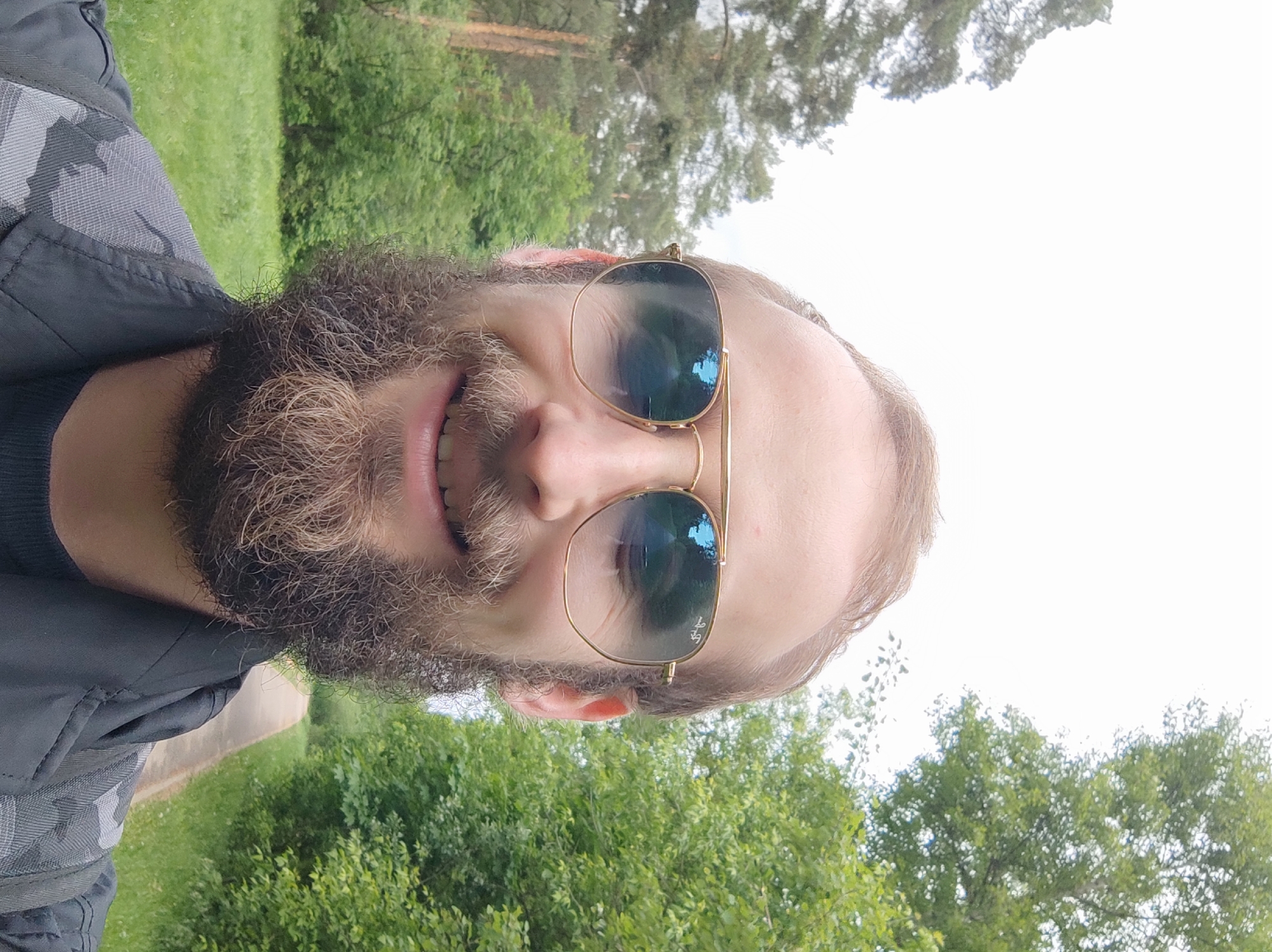 A selfie of a bearded man wearing gold-rimmed sunglasses. The background shows trees, suggesting a park or wooded area. A path or road is visible in the lower left corner. The location cannot be determined to be Moscow, Russia from the visual information provided.