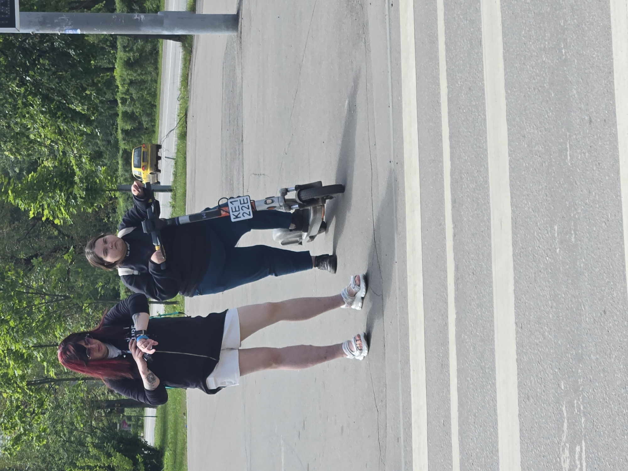 Two individuals are at a pedestrian crossing on a sunny day. The person on the left, with long red hair and glasses, is wearing a black zip-up jacket over a white shirt and white shorts, and is looking at a smartwatch on their left wrist. This individual has tattoos on their left forearm. They are wearing white sandals. The person on the right is on an electric scooter, wearing a black long-sleeved shirt and dark pants, and is looking forward. The electric scooter has a license plate that reads "KE1 22E". In the background, there is a yellow car on a road lined with trees and bushes. The location is indicated by street signs and the presence of a yellow car, suggesting a city environment. The overall scene appears to be in Moscow, Russia, as implied by the license plate format and general urban appearance.