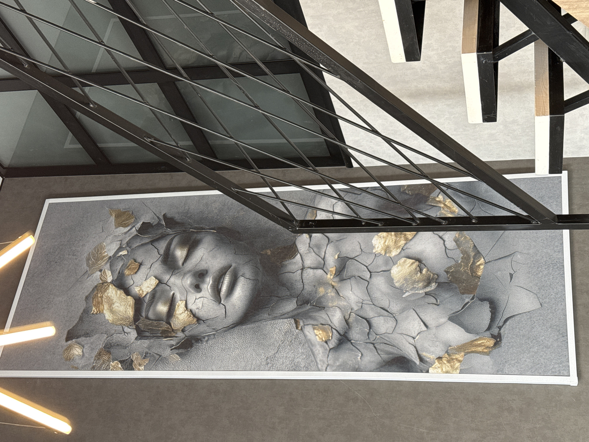 The image provides an overhead view of a contemporary indoor space, highlighting a distinctive piece of art. A large, rectangular framed artwork occupies the lower and central portion of the frame, depicting a stylized human face, likely female, rendered in grey tones with a cracked, textured appearance resembling stone or concrete. Gold metallic accents, resembling fractured pieces or torn leaves, are scattered across the surface of the face and its surrounding "cracked" skin, adding a contrasting, luminous element. The eyes of the face are closed, suggesting a serene or contemplative state.
Diagonally bisecting the image from top left to bottom right is a prominent dark metal structure, possibly part of a balustrade, staircase railing, or ceiling installation. It features a thicker main beam with several thinner, crisscrossing bars. This structure partially overlaps the top edge of the artwork.
In the upper right corner, beyond the metal framework, fragments of black and white furniture legs or architectural supports are visible against a light-toned, possibly textured, floor or wall. On the far left of the image, several modern linear light fixtures emit a bright, warm light, indicating artificial illumination. The floor in the foreground and bottom of the image is a solid, dark grey surface, possibly concrete or tile.
The scene is static, showcasing the interior design and artistic elements. The aesthetic combines industrial features with abstract art, suggesting a modern commercial or public environment such as a lobby, restaurant, or gallery. There is no visible activity or text.