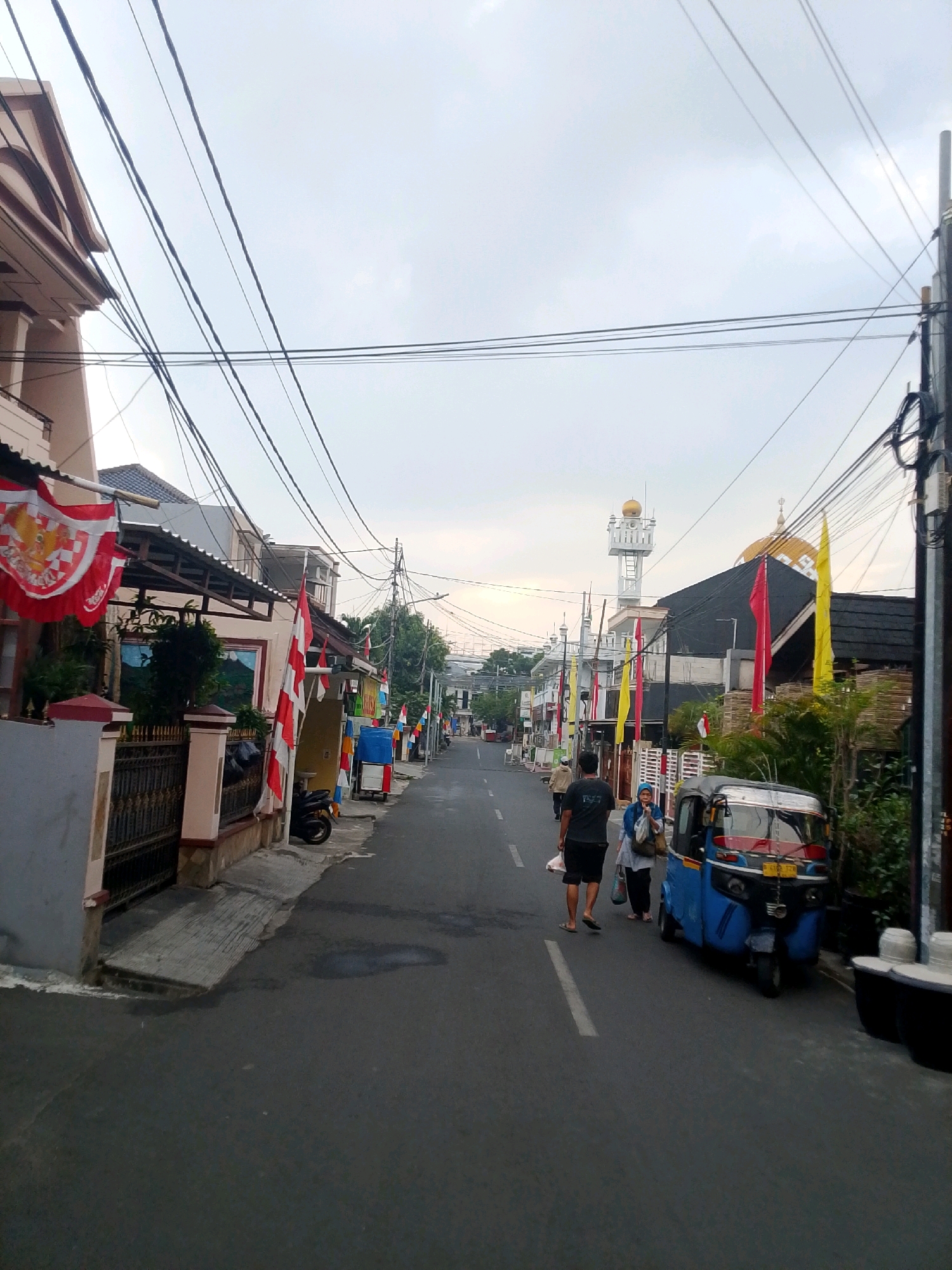 The image depicts a street scene in East Jakarta, Indonesia, likely during the early morning or late afternoon given the soft lighting and overcast sky. The street is paved and appears to be a residential or mixed-use area.
On the left side of the street, several houses are visible. One house in the foreground has a beige facade with arched details and is adorned with red and white flags, which are characteristic of Indonesian national celebrations. A black metal fence borders the property. Further down the street, more houses and buildings are visible, interspersed with trees. Indonesian flags, both small and large, are prominently displayed along the street and on buildings, suggesting a period of national observance.
On the right side of the street, a blue autorickshaw (bajaj) is parked. Two individuals are walking away from the camera on the right side of the street, with one person carrying bags. Another individual is walking further down the street. In the background, a mosque with a golden dome and a minaret is clearly visible, indicating the presence of a significant Muslim community in the area. Red and yellow flags are also flying near the mosque.
The sky is overcast, with a diffused light that casts soft shadows. Electrical wires are strung across the sky, a common sight in urban Indonesian environments. The overall impression is one of a bustling but serene street, decorated for a patriotic occasion.