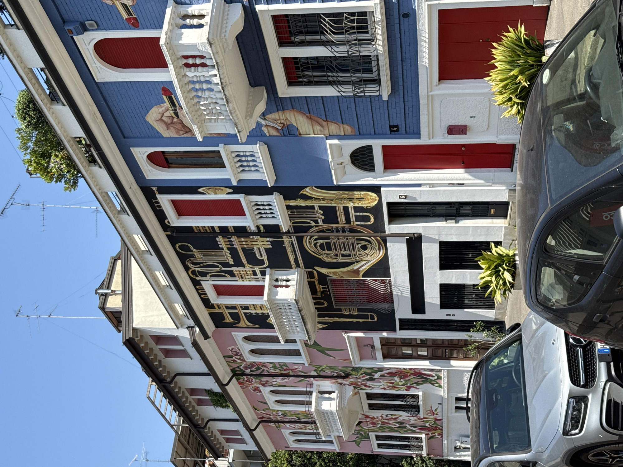 A low-angle, medium shot depicts the facades of colorful buildings in Milan, Italy. The buildings are three stories high and feature unique murals. The building on the left has a pink facade with a floral mural. The next building has a black facade with brass instruments painted on it. The building on the right has a blue facade with a hand holding a lipstick painted on it. All buildings have windows with white frames and red shutters. Cars are parked in front of the buildings. The sky is clear and blue.