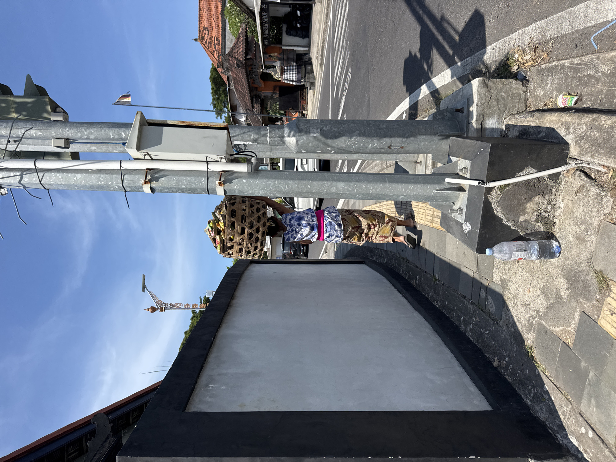On a bright, sunny day in Nusa Dua, Indonesia, an individual is seen carrying a woven basket laden with unidentified items, possibly offerings, atop their head. The person, dressed in a floral-patterned top and a brown patterned sarong with a pink sash, is walking along a concrete sidewalk. A metal street light post stands to the left, situated next to a white wall with a black trim. The background features a paved road, and buildings characteristic of Balinese architecture, including orange tile roofs and a barbershop. A partially visible German flag is attached to the street light. A discarded water bottle rests on the sidewalk near the wall.