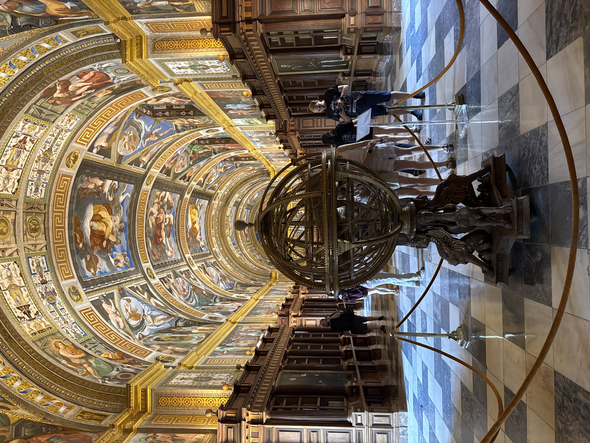 This image depicts the interior of the Royal Library of San Lorenzo de El Escorial, Spain, specifically the Hall of the Great Globe. The hall is characterized by its grand, arched ceiling adorned with intricate frescoes depicting biblical scenes and mythological figures, framed by decorative gold trim and Greek key patterns. The walls are lined with dark, ornate wooden bookshelves and display cases containing paintings, some of which are visible.
In the foreground, a large, intricately designed brass armillary sphere rests on a decorative pedestal featuring carved griffins and other mythical creatures. It is roped off with velvet ropes to prevent visitors from touching it.
Several people are present in the hall, appearing to be tourists or visitors. They are walking and observing the surroundings. One man in the middle ground is looking at his phone. Another group of individuals is standing near the armillary sphere, with one person pointing at an informational display. The floor is paved with a geometric pattern of light and dark marble tiles. The lighting appears to be natural, coming from unseen sources, and creates reflections on the polished floor. The overall atmosphere is one of historical grandeur and academic significance.