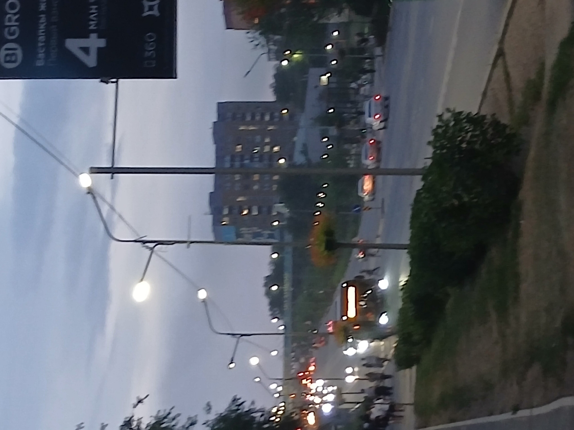 The image depicts a street scene in what appears to be a city during dusk or early evening. The sky is overcast and a pale blue-grey. Streetlights are illuminated, casting a glow on the asphalt road and sidewalks.
In the foreground, a grassy verge with a low hedge runs along the left side of the image. A sidewalk borders this verge.
The main focus of the scene is the road, where several vehicles are present. A bus with its headlights and interior lights on is prominently positioned in the center, moving away from the viewer. To the right of the bus, a white car is visible. Further down the road, two cars with their brake lights illuminated are driving away.
On the sidewalks, groups of pedestrians are visible. On the left side, a cluster of people are walking along the sidewalk. On the right side, near the bus, more figures are dispersed along the sidewalk.
In the background, a large, multi-story building with numerous lit windows stands out. Trees and other vegetation are interspersed throughout the scene, some illuminated by the streetlights.
A dark sign is mounted on a pole on the right side of the image, displaying text and a large number "4". Another smaller sign with "0360" is also visible.