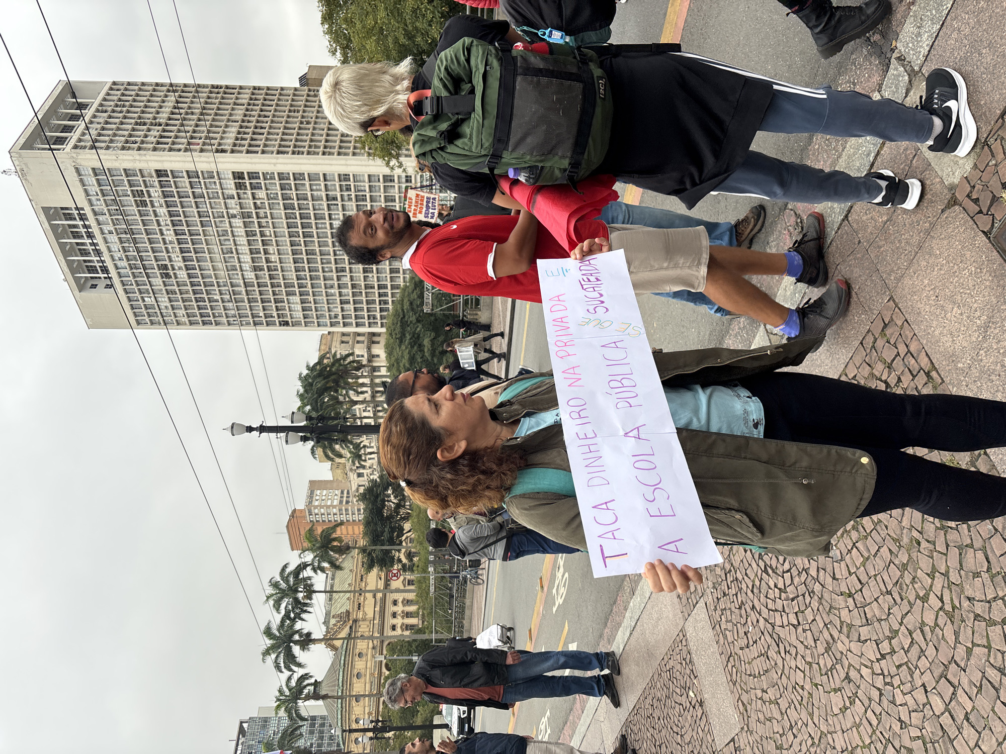 The image captures a protest in the city of São Paulo, Brazil. A woman stands in the foreground, holding a white sign with text written in Portuguese, advocating for public schools and against privatization. She is wearing a green jacket and dark pants. Behind her, other people are seen protesting, including a man in a red shirt and beige shorts holding a similar sign. In the background, there's a view of the city, with buildings, palm trees, and light poles visible. The overall atmosphere suggests a public demonstration or rally.