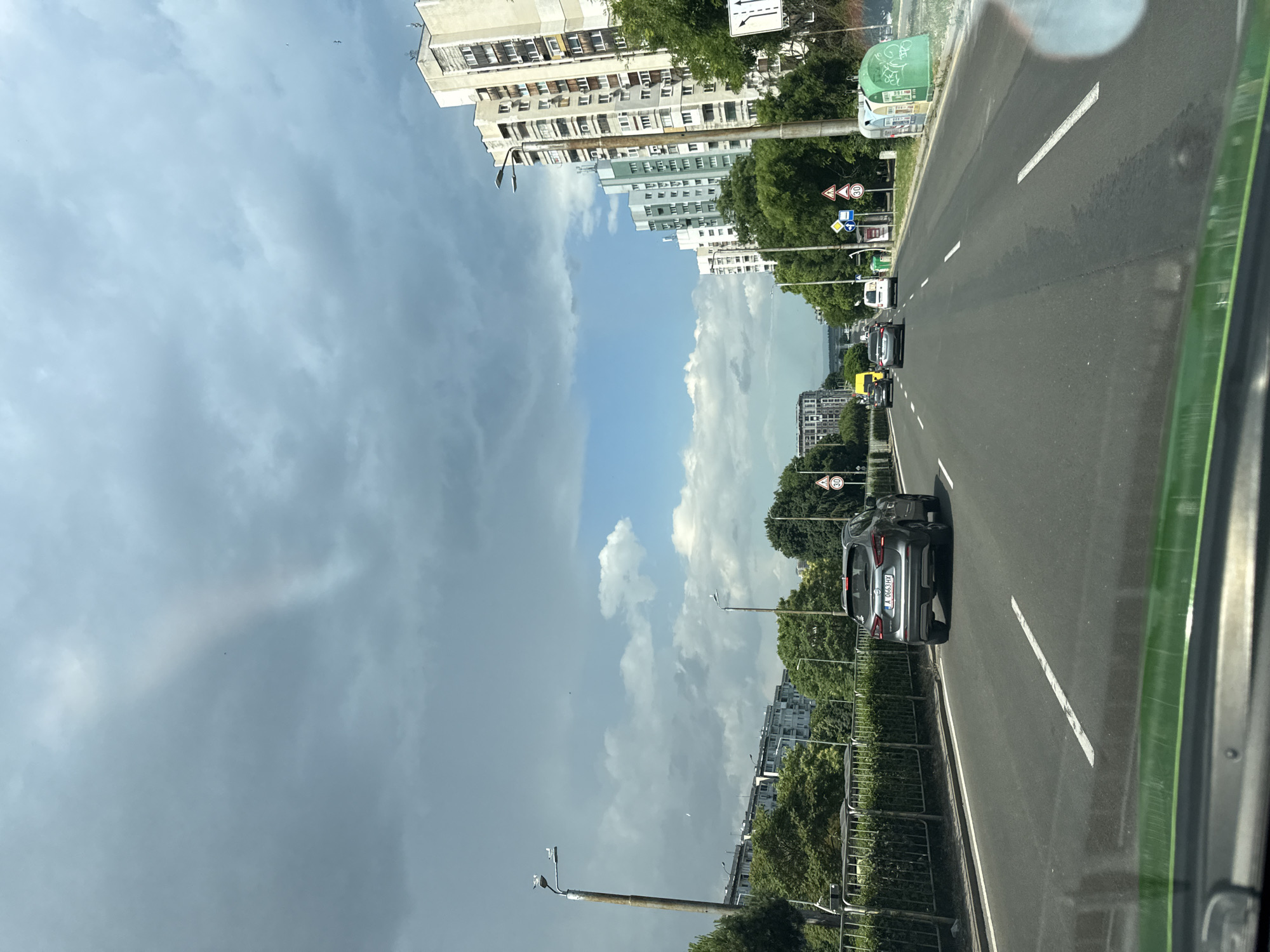 This media file captures a street scene in Burgas, Bulgaria, viewed from inside a vehicle. The perspective is unusual, with the image rotated 90 degrees counter-clockwise. The foreground is dominated by the car's interior, with a green frame visible on the right.
The scene depicts a multi-lane road with several cars driving in the same direction. A dark grey car with a license plate starting with "A 965 HX" is prominently featured. On the left side of the road, lined with trees, are several buildings, some appearing to be apartment blocks with many windows. There are also streetlights and traffic signs, including speed limit signs displaying "30" and road directional signs.
The sky is a mix of blue with scattered white clouds and darker, stormy-looking clouds, suggesting a dynamic weather pattern. The time of day appears to be daytime, given the natural light illuminating the scene. The overall impression is of a typical urban environment on a day with changing weather conditions. The rotation of the image creates a surreal effect, making the sky appear to be on the left side of the frame and the road and buildings on the right. No people are clearly identifiable as subjects.