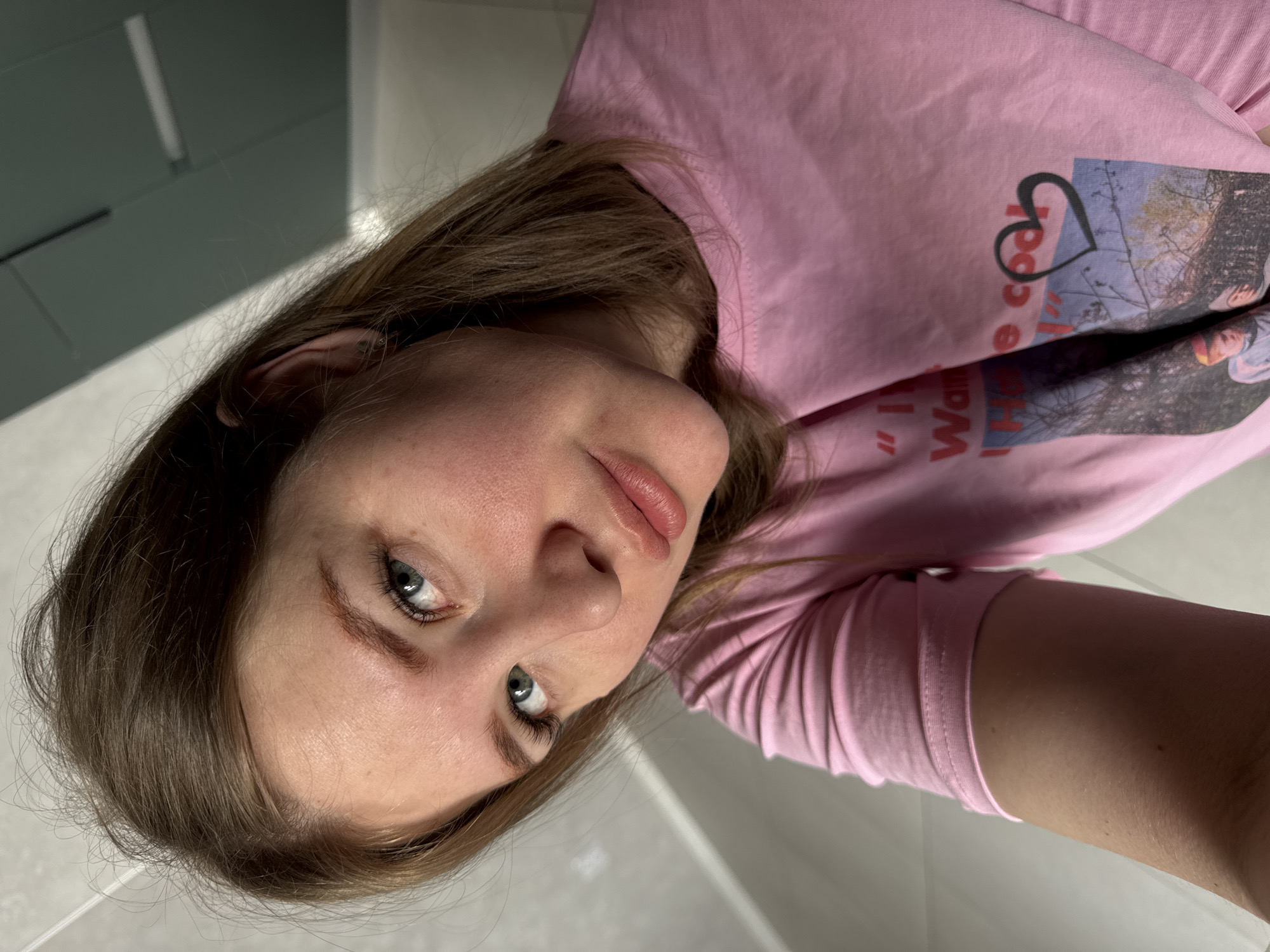 An indoor photograph, located in Gornyi (Dvurechenskoe S/P), Russia, presents a female subject positioned from the mid-torso upwards, angled slightly downwards into the camera lens. The subject has light brown hair, blue-green eyes, and a fair complexion with visible redness on the nose and cheeks. She wears a pink short-sleeved t-shirt featuring a partial graphic print on the front, which includes fragmented text, an outdoor scene with trees, and a black heart outline. Her expression is neutral, and her gaze is directed at the camera. The background consists of light-colored, plain walls, with subtle horizontal lines visible towards the top of the frame, indicating internal building structures or paneling.