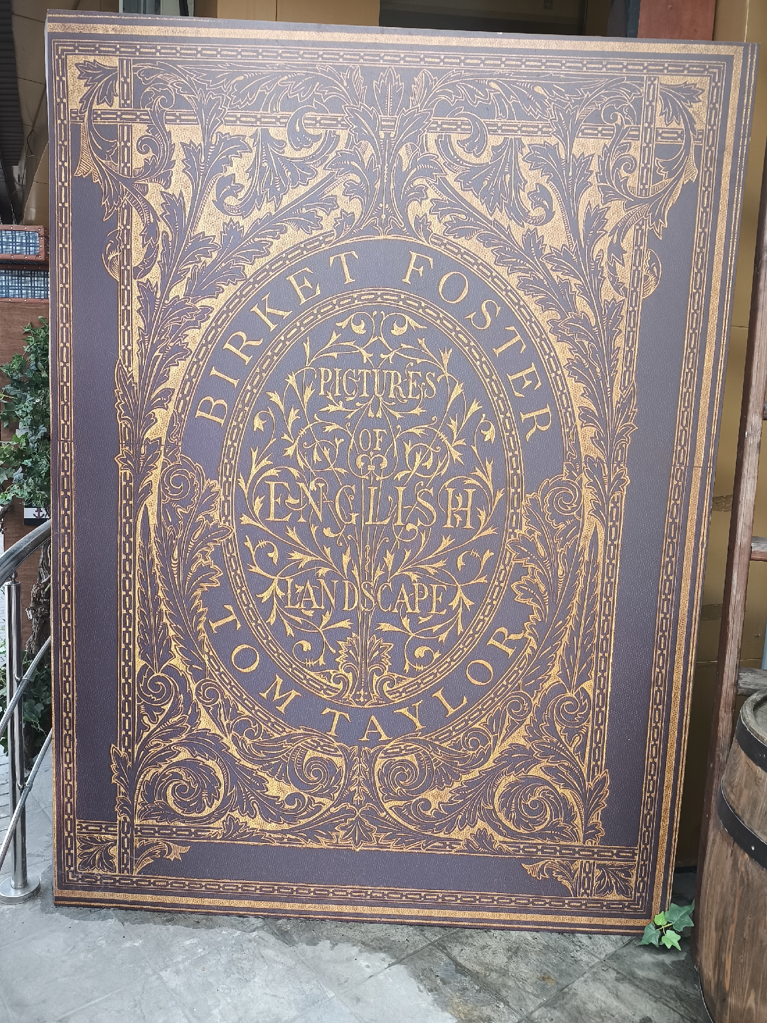 A large, rectangular sign is leaning against a wall or structure outdoors. The sign features an elaborate design reminiscent of an old book cover. The color scheme is dominated by a dark purple or mauve background with intricate gold detailing. The text "BIRKET FOSTER" arches across the top of an oval design. Below this, the words "PICTURES OF ENGLISH LANDSCAPE" are arranged in a stacked formation within the oval. At the bottom of the oval, the text "TOM TAYLOR" arches similarly to the top text. The decorative elements include floral and vine-like patterns.
The sign appears to be situated in a public space, likely a street or pedestrian area. To the left, a metal railing and some greenery are visible. To the right, a wooden barrel is partially visible. The ground is paved, and a small plant is growing near the base of the sign.
Based on the architectural style and information within the image, it is likely this photograph was not taken in Moscow, Russia.