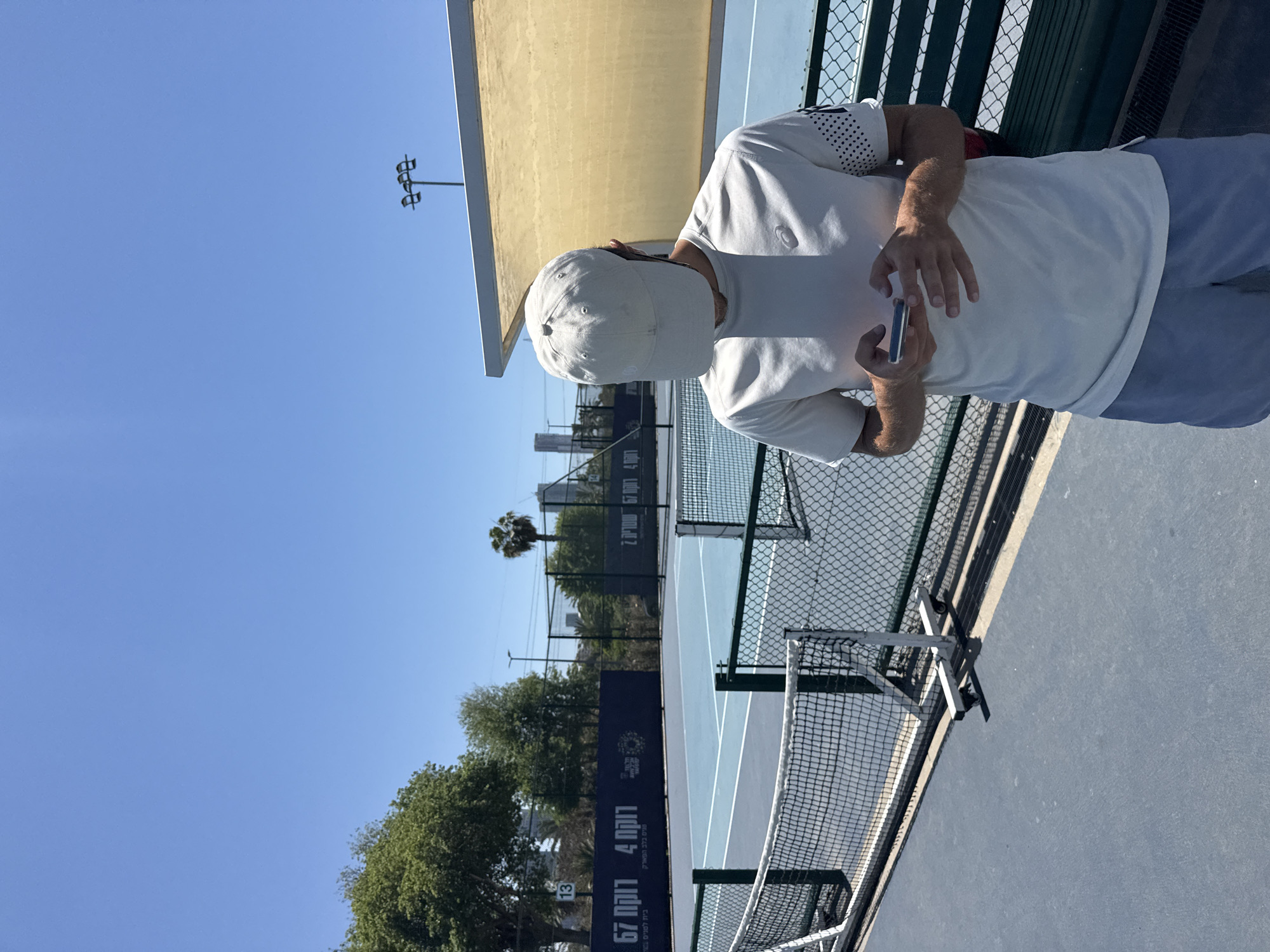 The image depicts a man seated on a bench next to a blue padel tennis court in Tel Aviv, Israel. He is wearing a white cap, a white t-shirt with a patterned sleeve, and light-colored pants. He is holding a mobile phone in his hands, looking at it. The tennis court is enclosed by a green fence and net. Behind the court, there are trees and a tall palm tree visible in the background. A yellow building is on the left side of the image. The sky is clear blue. A light post is visible in the background. A sign in Hebrew is adjacent to the court.