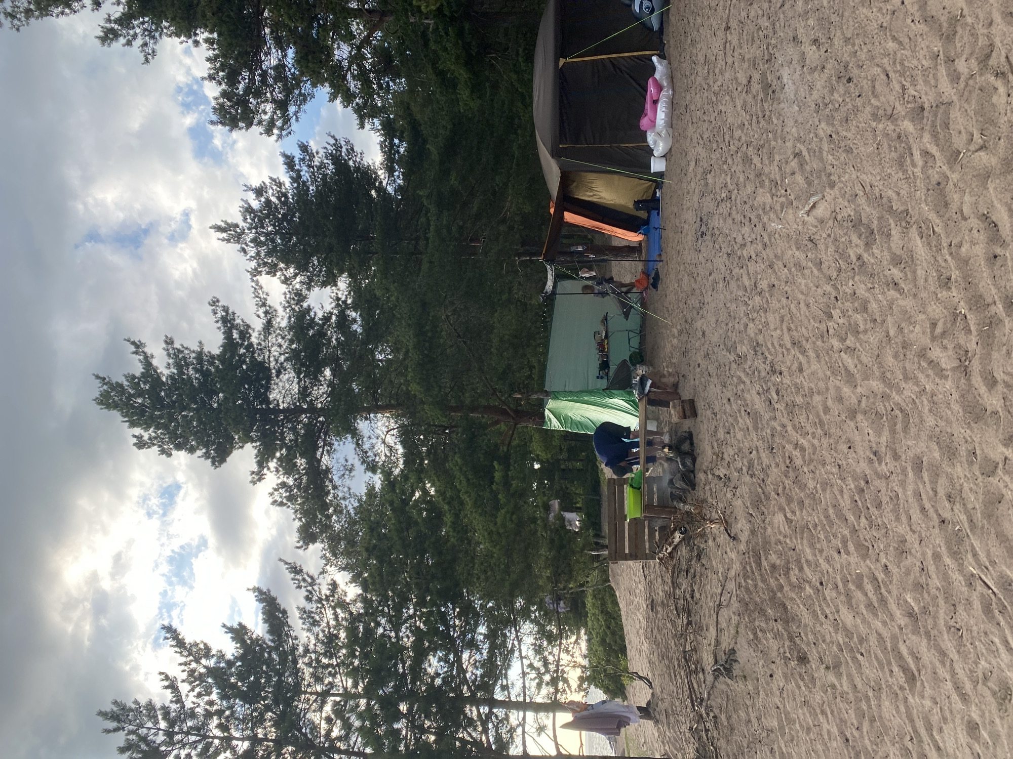 The image depicts a sandy campsite near a body of water, bordered by a dense pine forest. The sky is partly cloudy with patches of sunlight breaking through. In the foreground, the sandy ground is textured with ripples from the wind.
Several tents are set up on the sand. A large, dark-colored tent is prominent on the right side of the frame, with an orange-colored awning. Next to it, a smaller, lighter-colored tent is visible, partially obscured. To the left, a green tarp is stretched between poles, creating a covered area. Underneath this tarp, a table is set up with various items, including what appears to be a green basin and a person wearing a dark blue jacket and shorts, interacting with something on the table.
In the middle ground, a wooden structure resembling a picnic table or bench is positioned near a small campfire with visible embers. A bucket with a green lid is on the table. To the left of this structure, a person in a white garment is standing near a pole where several items of clothing are hanging to dry.
The environment suggests a natural setting, likely a beach or shoreline, with the Baltic Sea possibly in the background, indicated by the presence of water and the direction of the sunlight. The presence of tents, a campfire, and drying clothes suggests a recreational camping activity. The location is identified as Baltiiskoe, Russia, a coastal area.