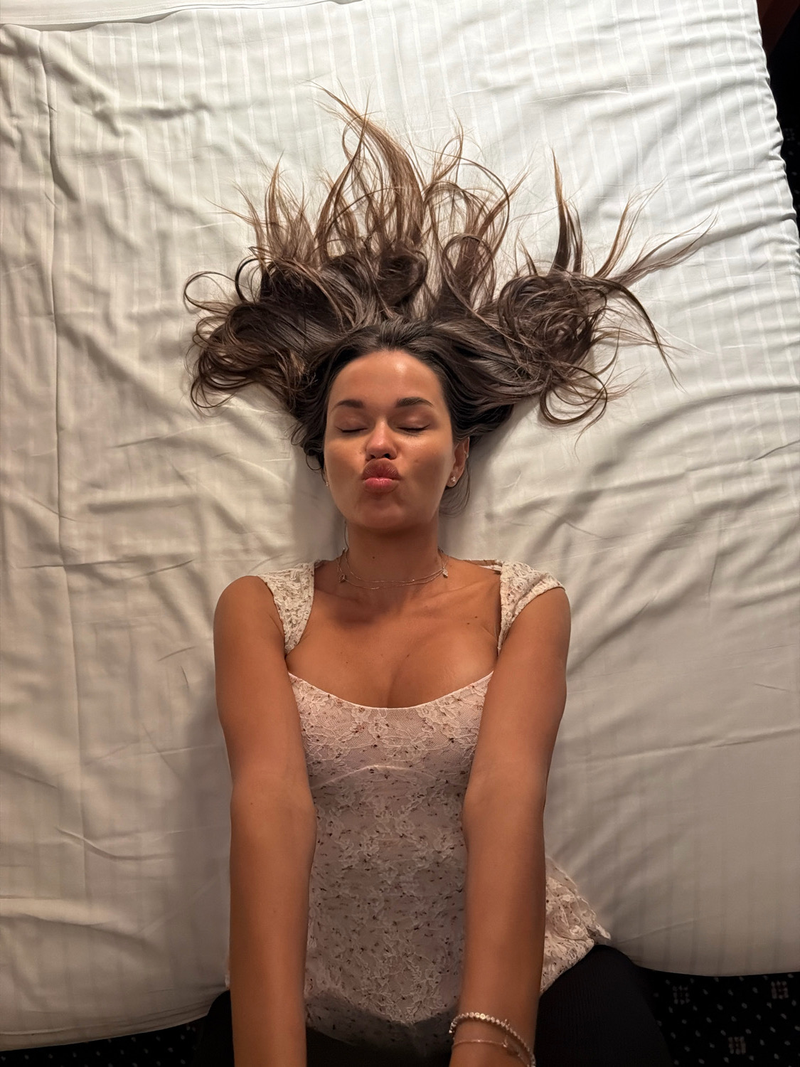 A young woman with long, dark brown hair lies on her back on a white, striped bedsheet, her eyes closed and lips pursed in a kiss. Her hair is spread out around her head. She is wearing a floral print, lace-trimmed top and a thin silver necklace. Her arms are bent at the elbows and resting on her chest. The scene is indoors, on a bed. There is no indication of the location in Verona, Italy.