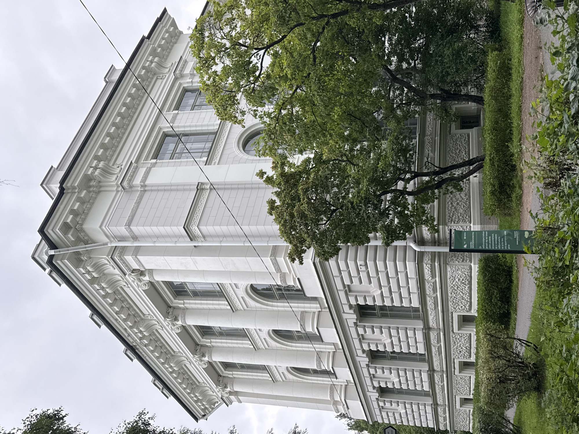 A low-angle, full-shot photograph captures the exterior of a large, white, multi-story building with classical architectural elements. The building features columns, arched windows, and ornate detailing along its facade. Trees with lush green foliage partially obscure the view of the building, with branches and leaves extending into the frame. The sky above is overcast and gray. In the foreground, a paved walkway is visible, bordered by green bushes and grass. A dark green informational sign with white text is positioned on the right side of the walkway. The scene suggests an outdoor setting in a city, likely during daylight hours with muted natural light. The location is specified as Ladozhskoe Ozero, Russia. There are no people visible in the image.