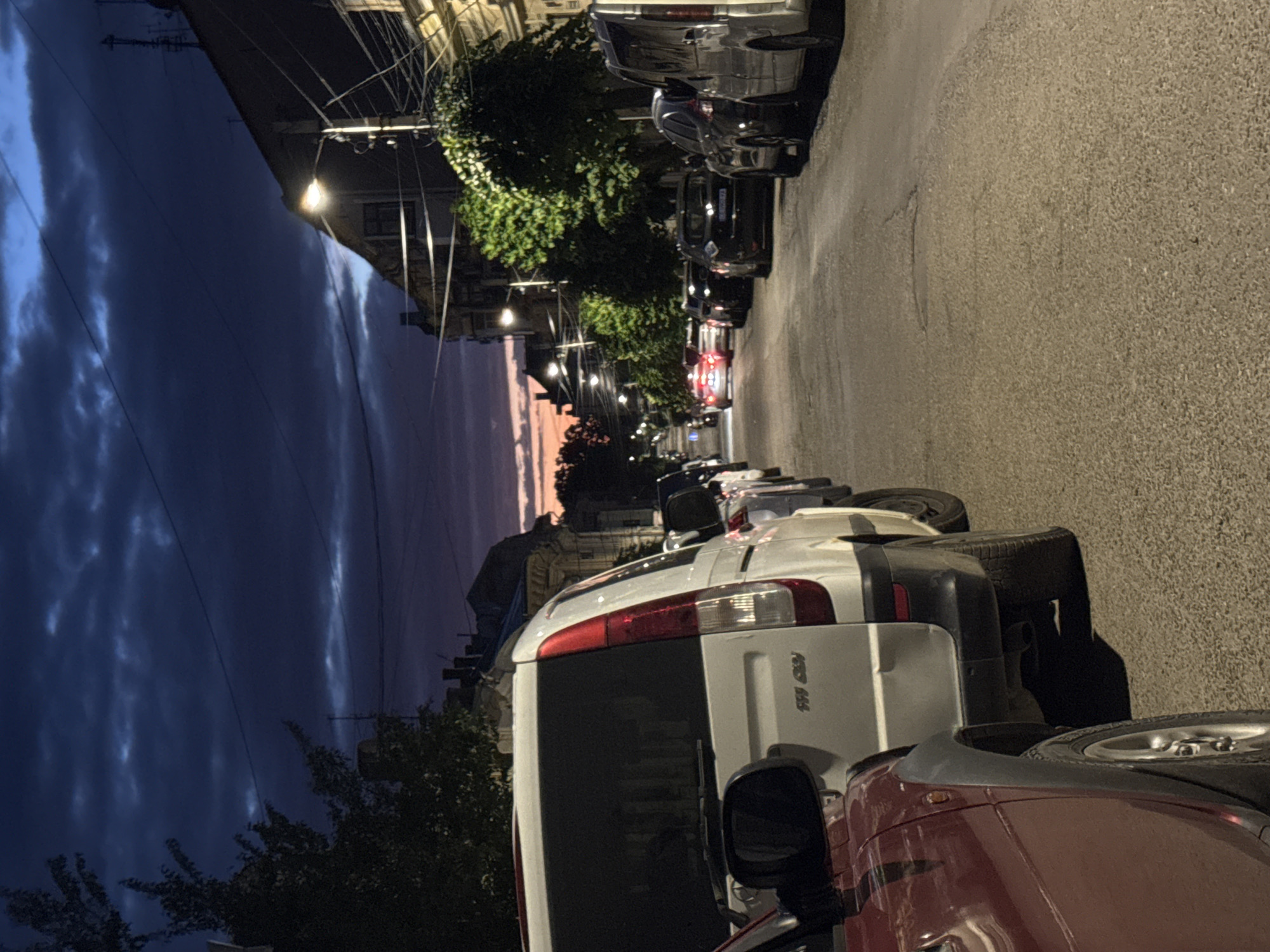 The image depicts a quiet street in Chernivtsi, Ukraine, at dusk or early night. The sky is a deep blue with hints of a sunset visible in the distance. Several cars are parked along the street, creating a sense of stillness and routine. The architecture of the buildings lining the street suggests an older urban environment, with some architectural details hinting at historical influences. The streetlights are illuminated, casting a warm glow on the asphalt and the parked vehicles. The overall atmosphere is peaceful and ordinary, capturing a moment of urban tranquility as day transitions into night.
