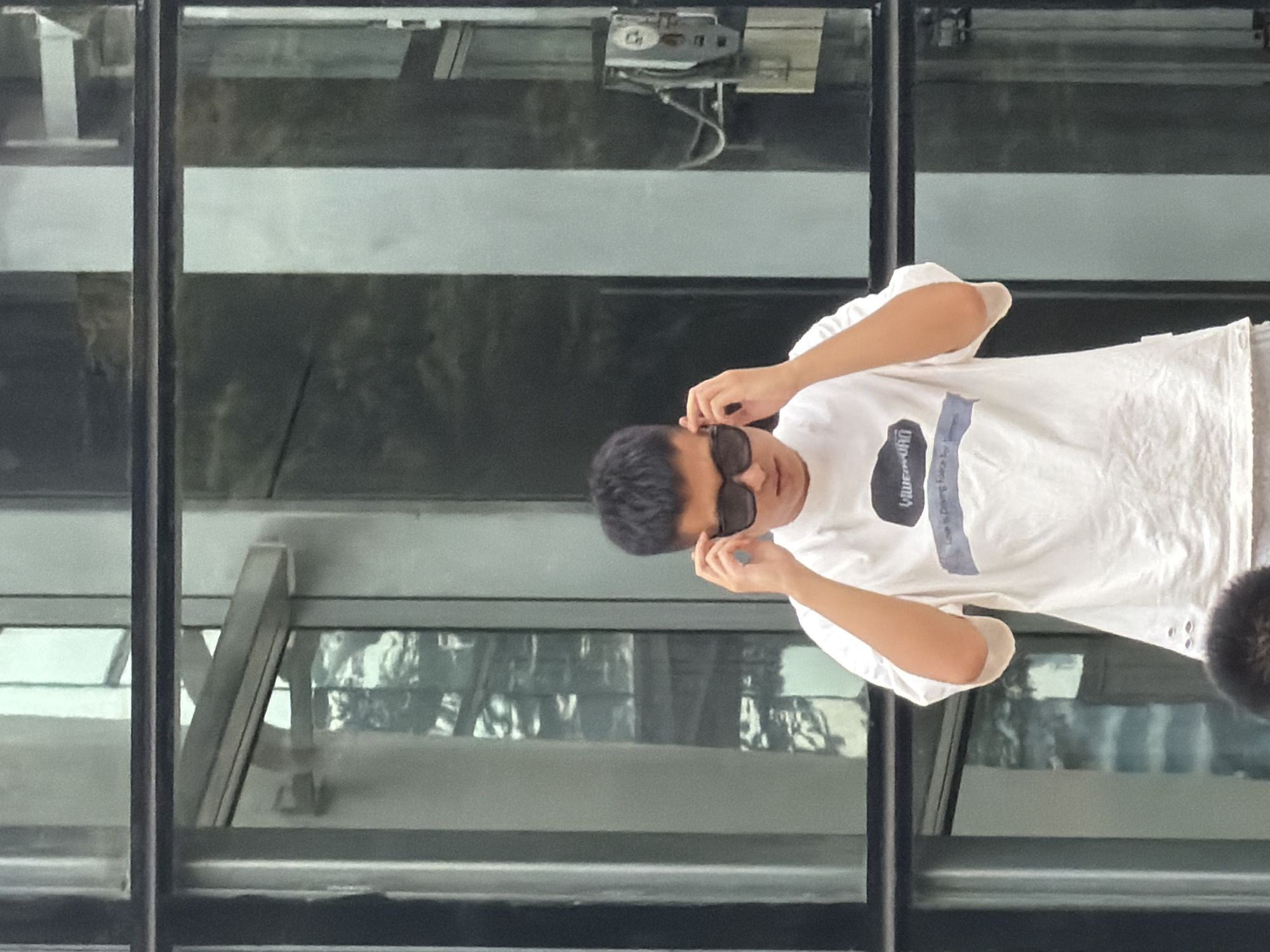 In Yuzhong District, China, a young East Asian male stands in front of a building with large glass windows, which reflect the surrounding environment. He is wearing a white t-shirt with a graphic print and light-colored shorts. The male is adjusting his dark sunglasses, with his hands near his temples. The background appears to be a modern architectural structure, with a metallic frame and glass panels. The lighting suggests it is daytime, possibly overcast, as there are no strong shadows. A grey electrical box is mounted on the wall to the right of the subject.
