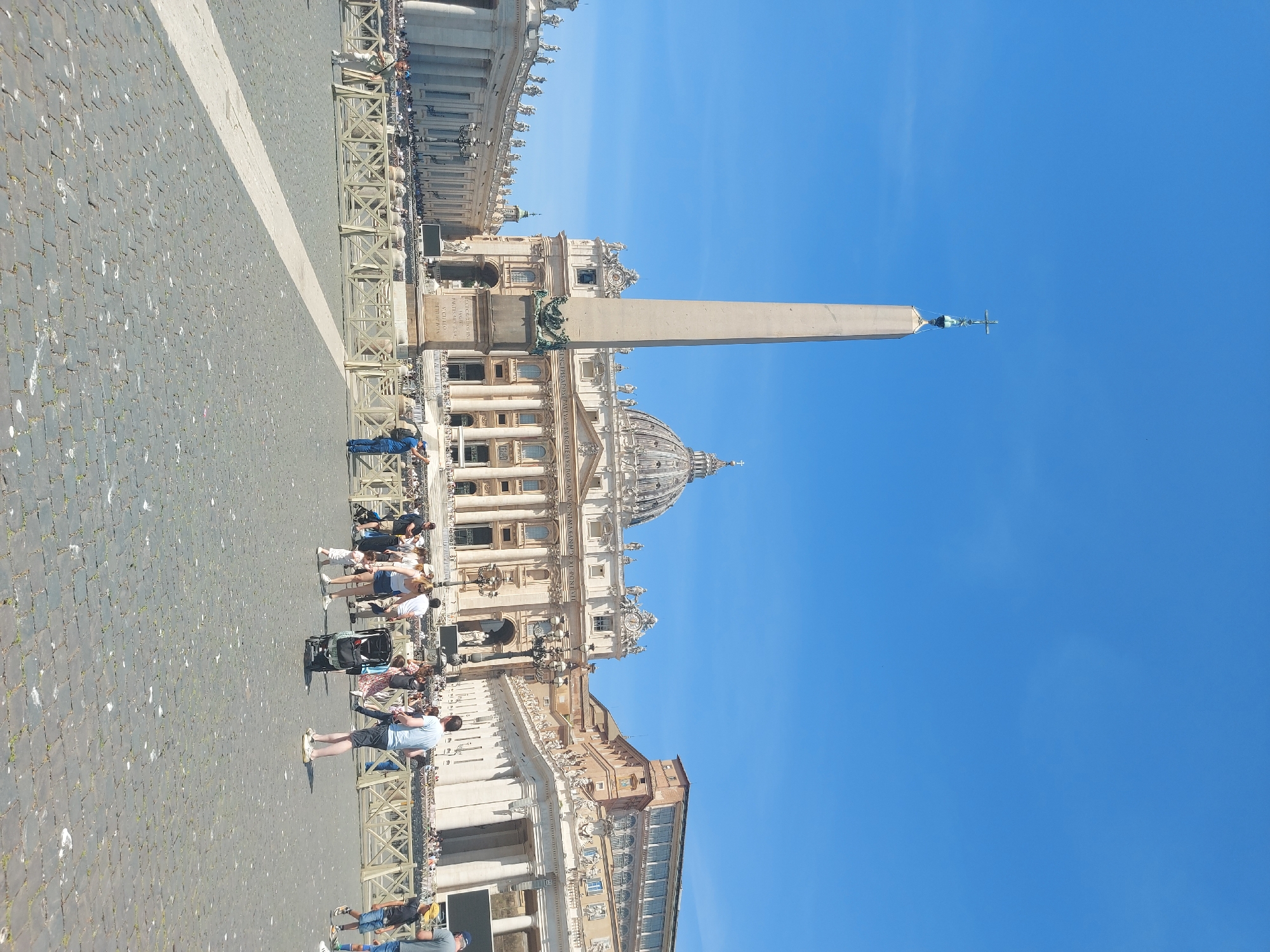 The image depicts Saint Peter's Square in Vatican City, Roma, Italy. The square is paved with cobblestones and features an obelisk in the center. St. Peter's Basilica stands prominently in the background with its dome visible. People are seen walking and standing in the square. A low barrier is visible along the perimeter. The sky is clear and blue.