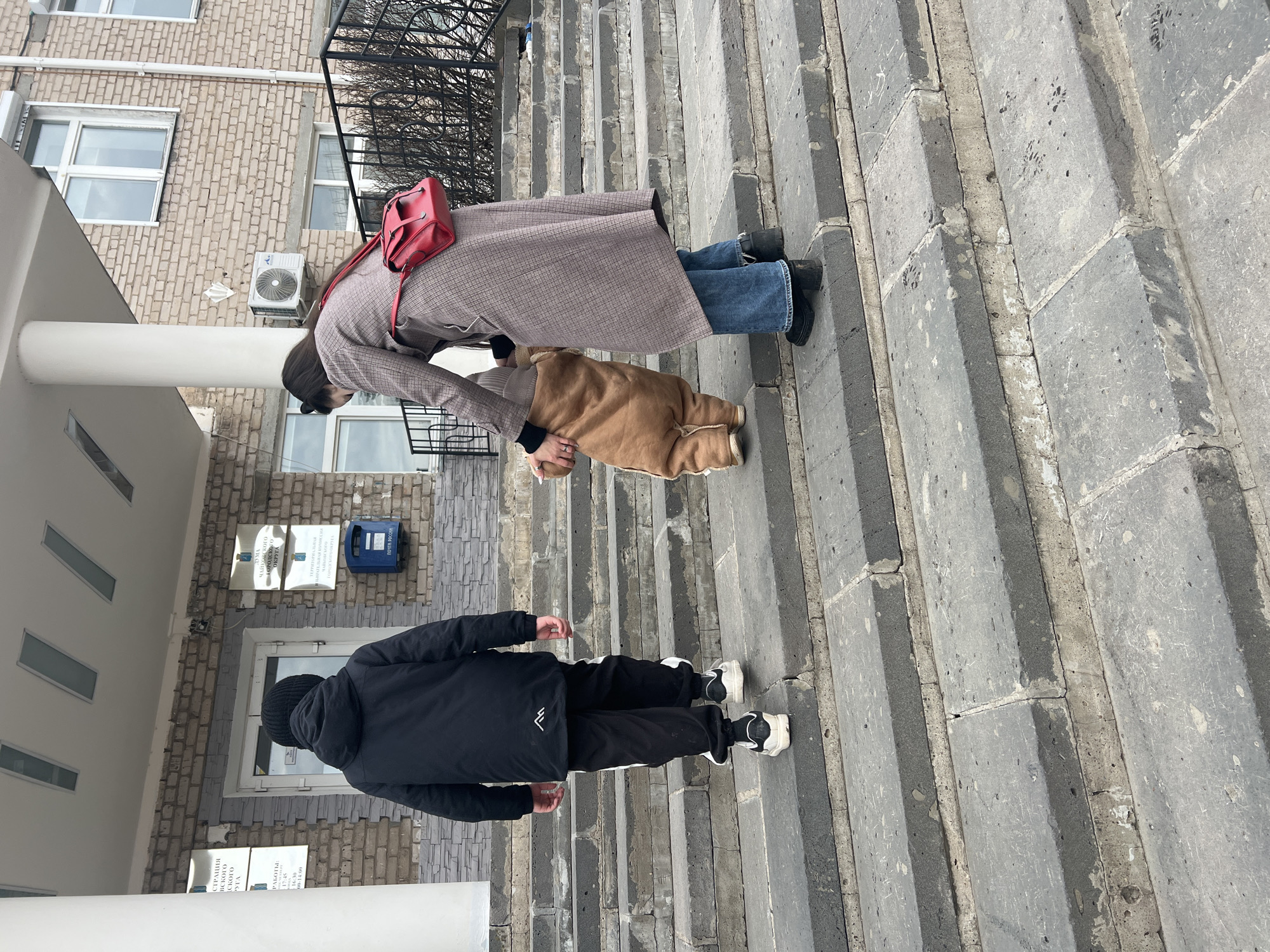 The image shows two people ascending a set of concrete stairs leading to a building in Chaikovsky, Russia. One person, possibly a woman, is wearing a plaid coat, blue jeans, and a red backpack. They are bent over, seemingly attending to a young child clad in a brown jacket. Another person, dressed in a black jacket and pants with white stripes, is further down the stairs, walking upwards. The building is made of light-colored brick and features a mailbox and informational signs near the entrance. There are windows and an air conditioning unit visible on the building's exterior.